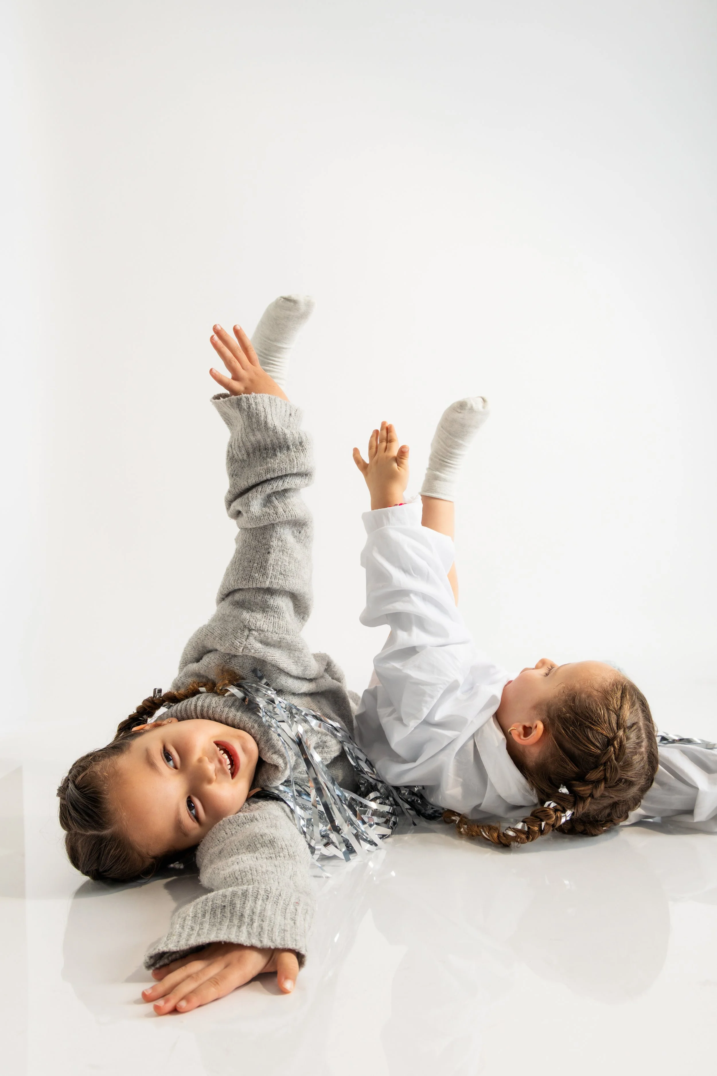 Two children with braided hair lying on the floor. One girl with gray sweater and metallic tinsel around her neck, smiling at the camera. Another girl in a white shirt and gray socks, lying on her back with arms raised, looking sideways. Both are aga