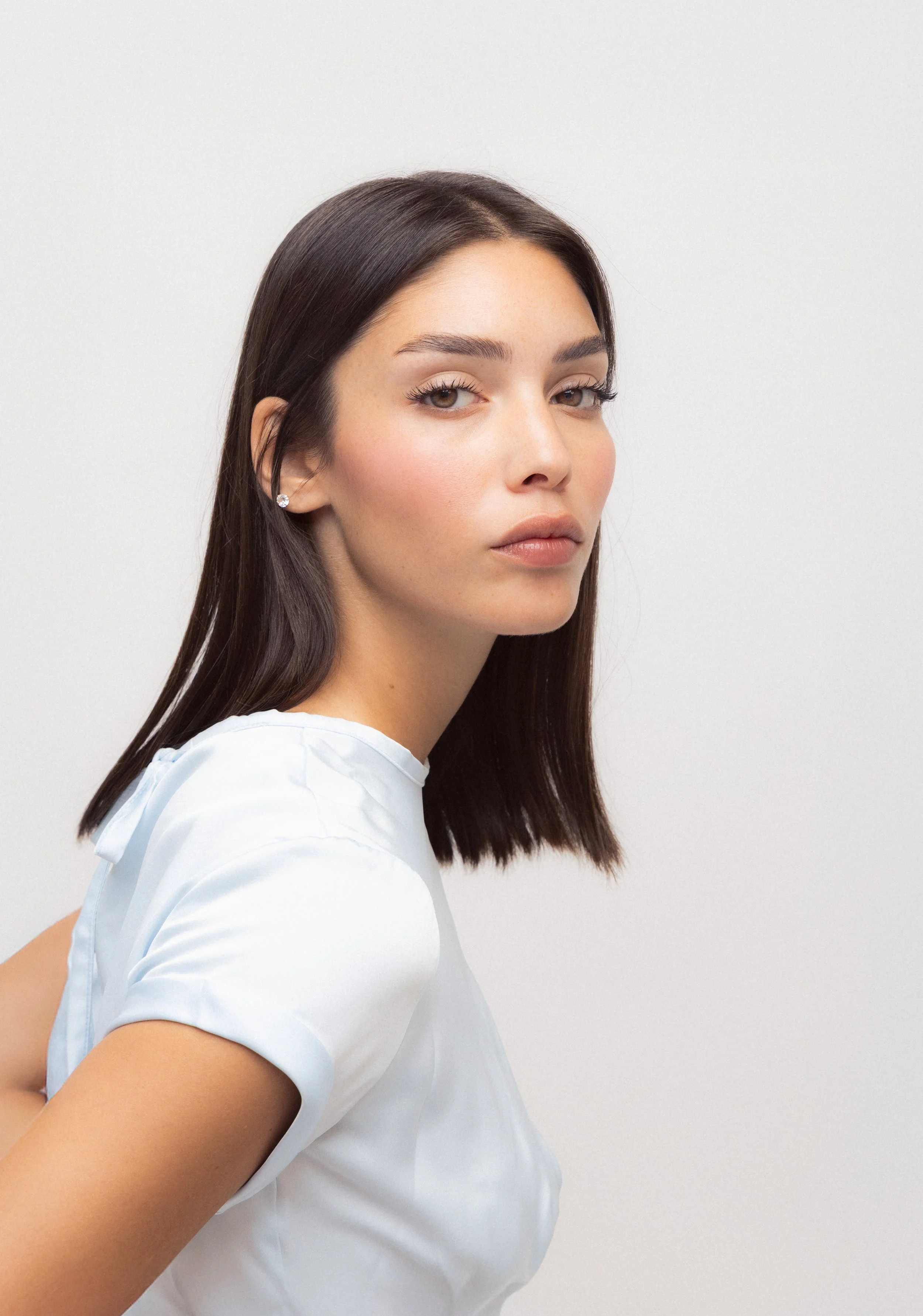 A woman with long dark hair and light makeup wearing a white top against a plain light gray background.