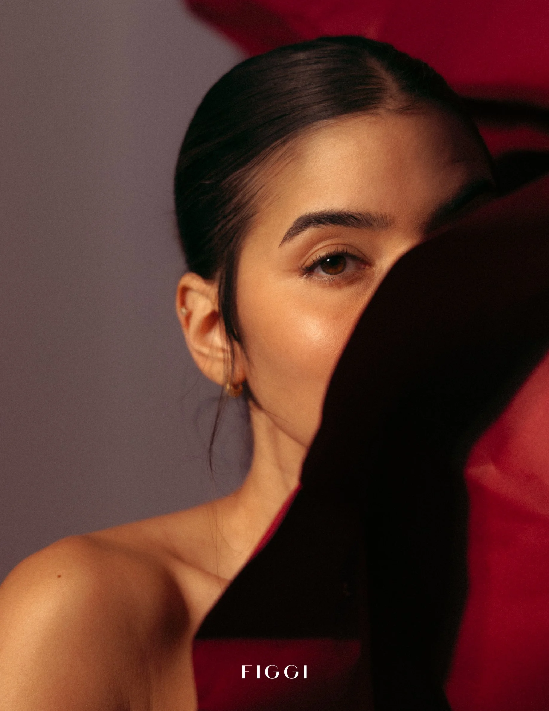 Close-up of a woman's partially visible face, showing her eye, eyebrow, and part of her cheek, with dark hair pulled back, against a red background.