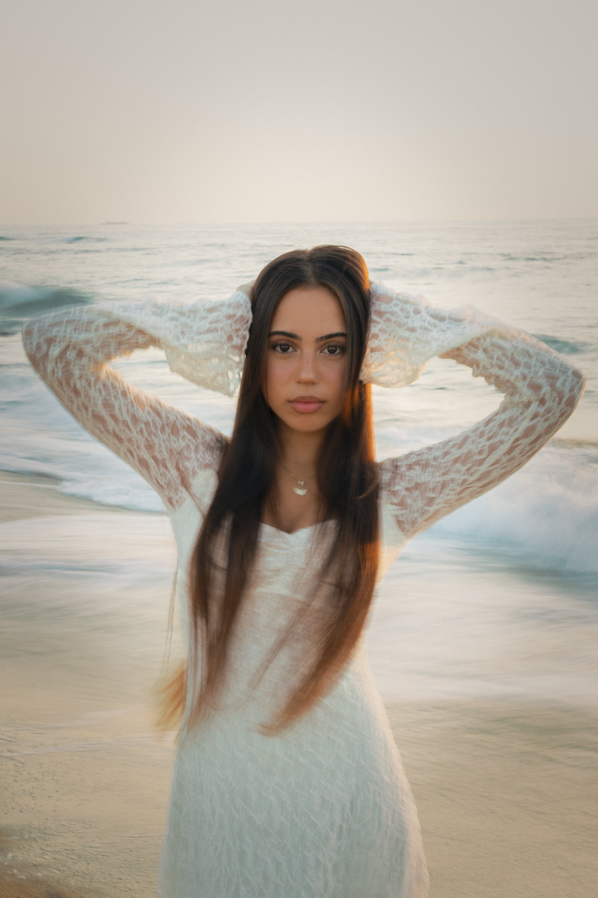 A young woman with long brown hair and a white lace dress standing on a beach at sunset, with the ocean and sky in the background.