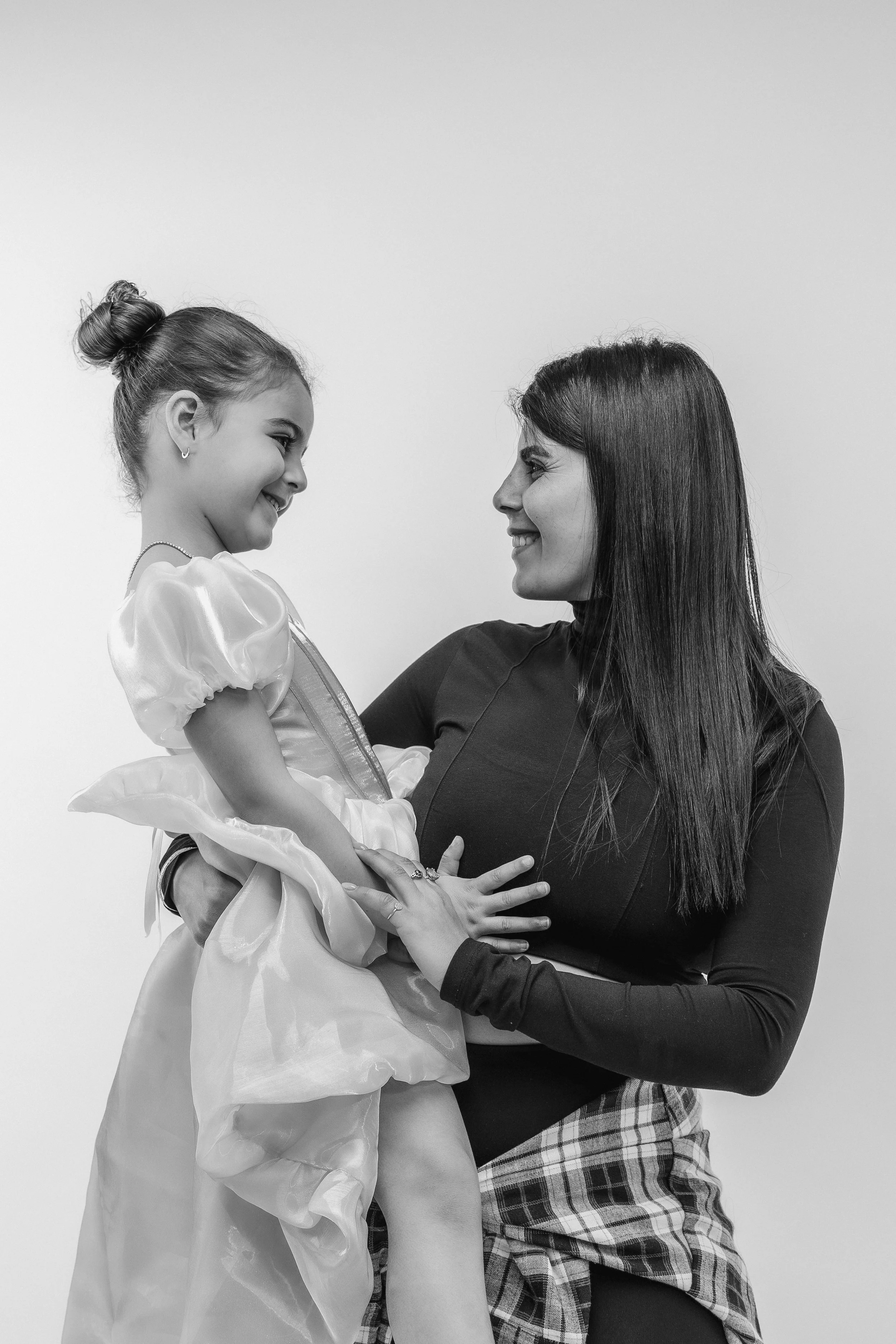 A woman with long dark hair holding a young girl in a princess costume, both smiling at each other, black and white photograph.