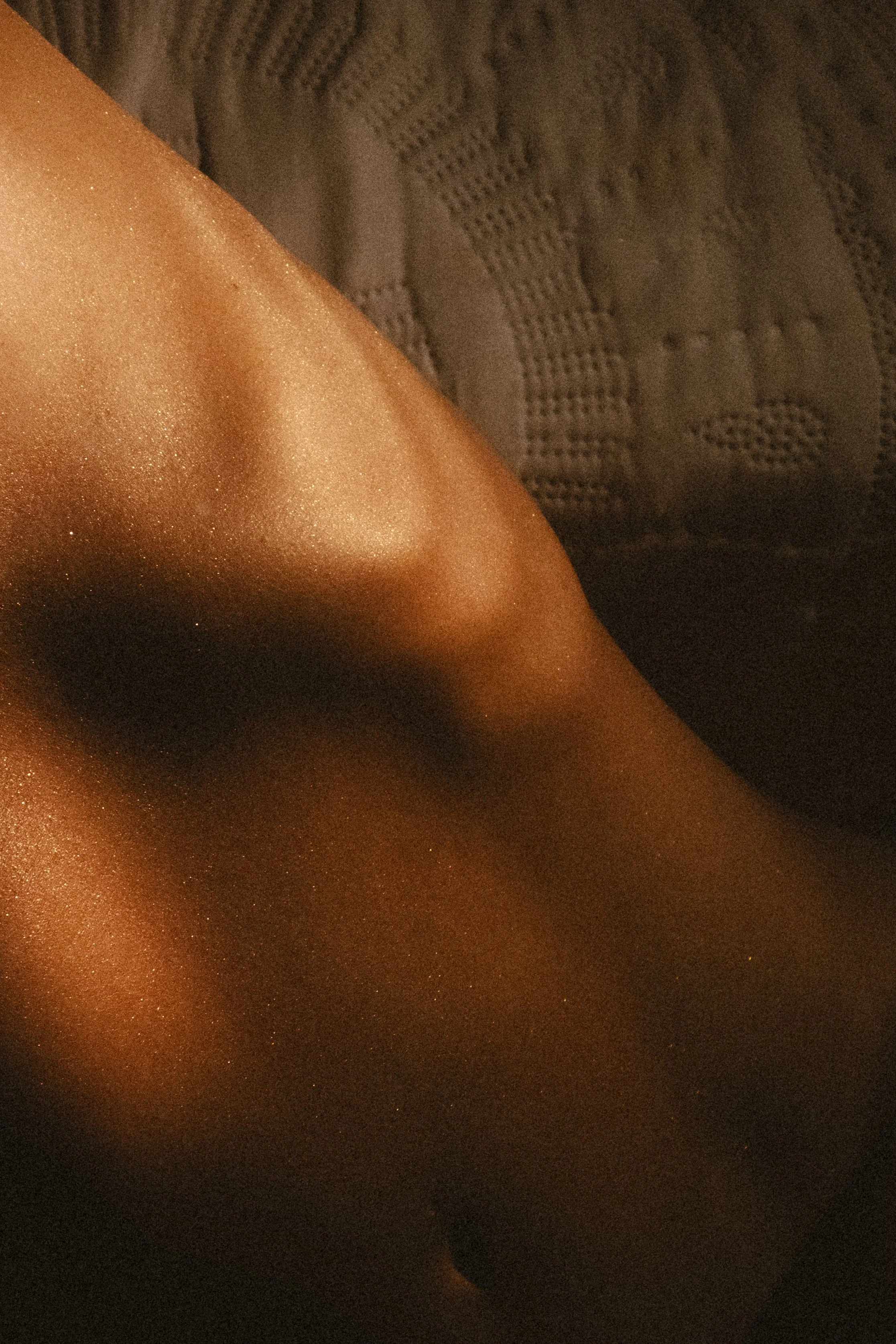 Close-up image of a person's arm with a metallic or shimmery effect, possibly from body lotion or makeup, against a dark textured background.