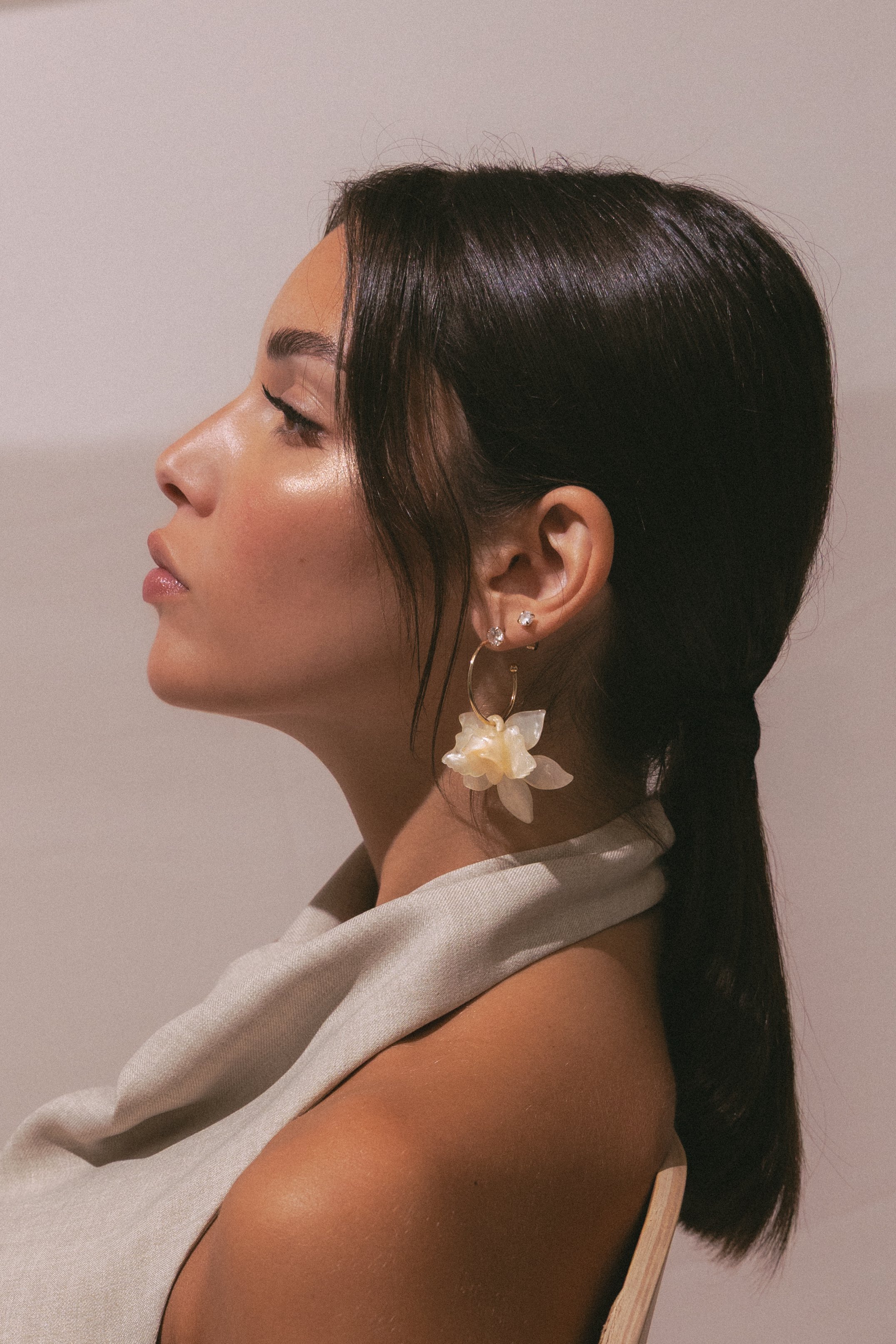 Profile of a woman with dark brown hair styled in a bun, wearing large flower-shaped earrings, a single pearl earring, and a beige satin outfit, against a plain light-colored background.