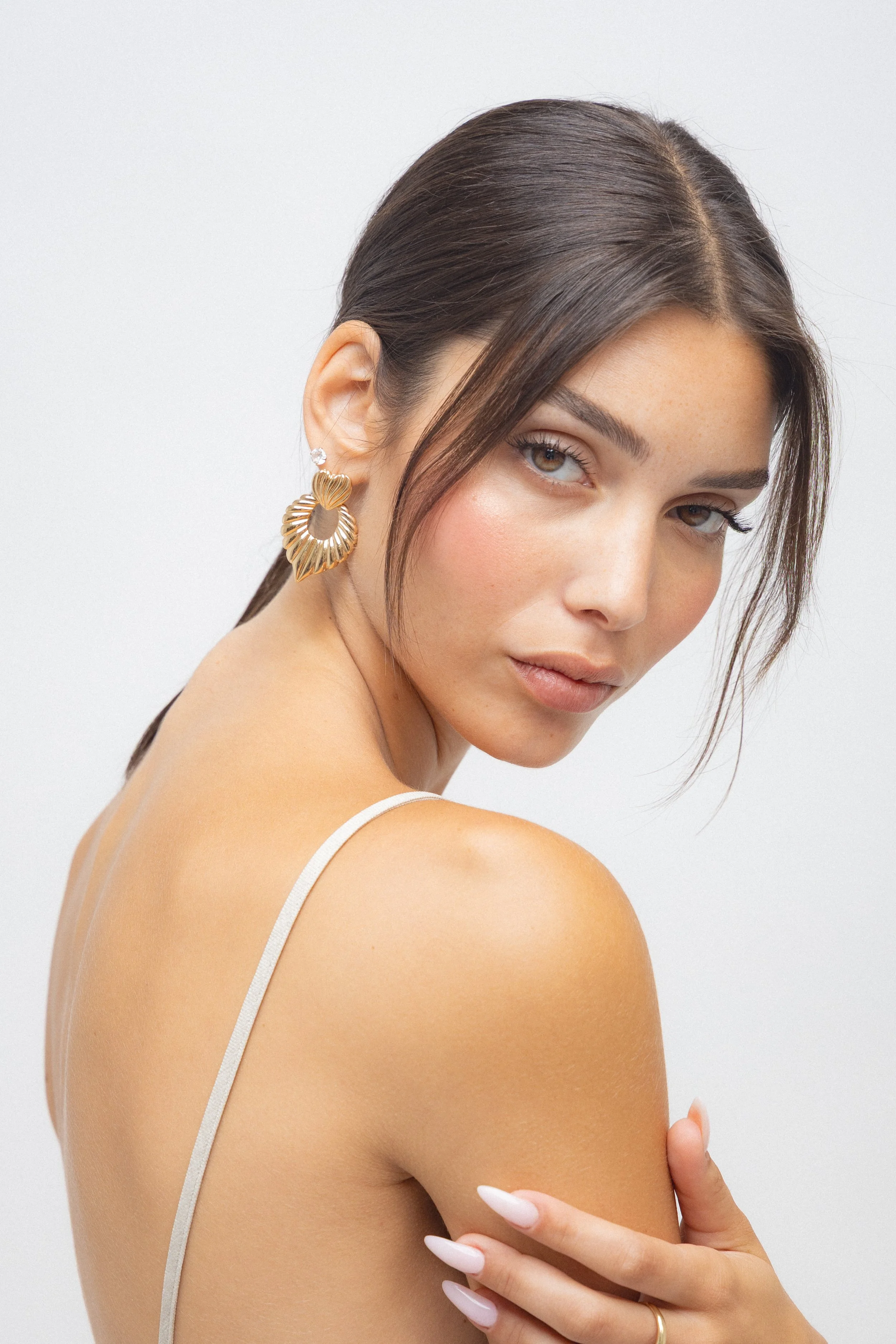 Close-up of a woman with dark brown hair styled back, wearing large gold heart-shaped earrings, lightly touching her shoulder, with a soft expression and natural makeup, against a plain white background.