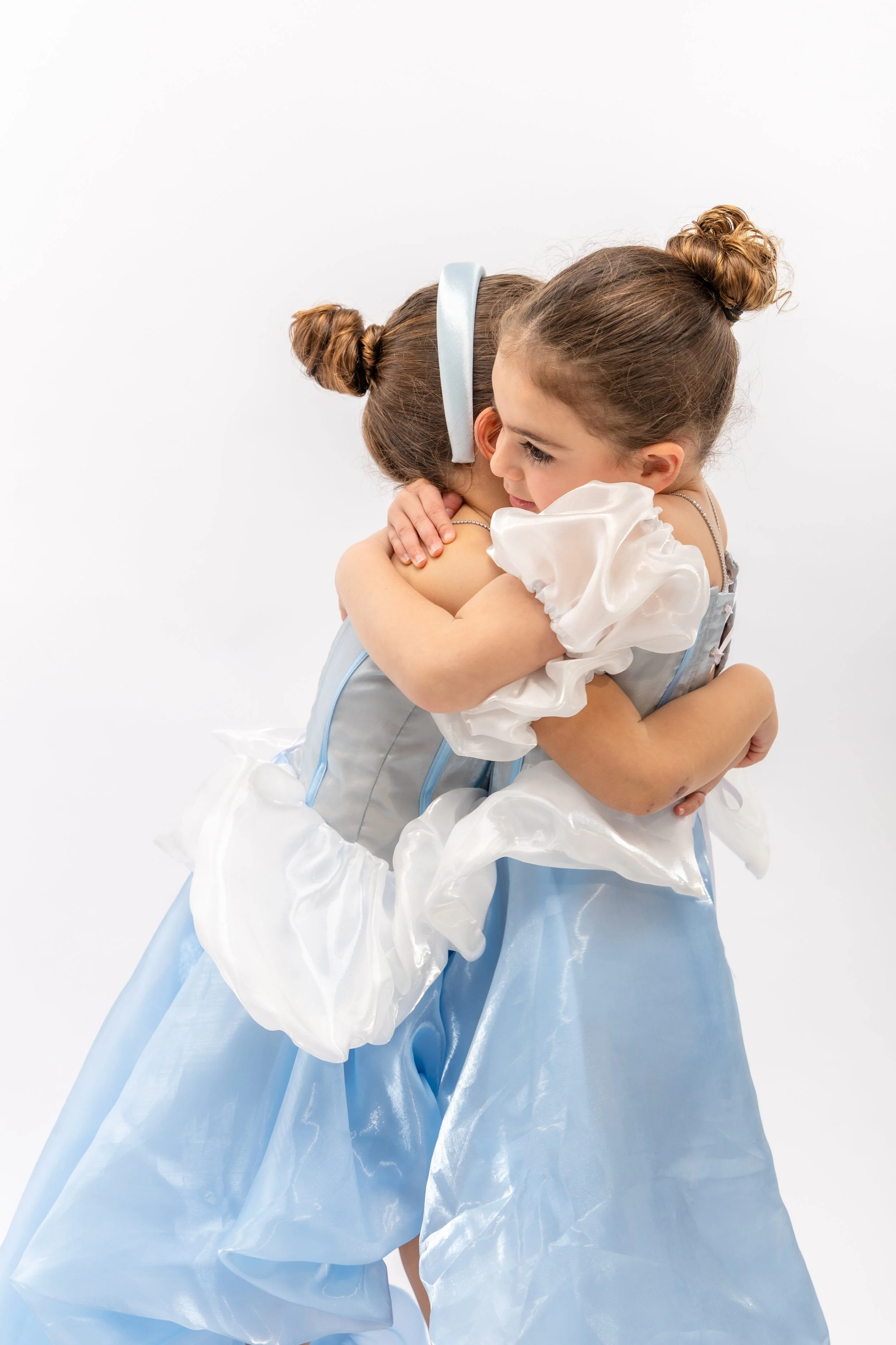 Two young girls hugging each other, one dressed as a princess with a light blue gown and the other wearing a white dress with puffy sleeves, both with their hair styled in buns, against a plain white background.