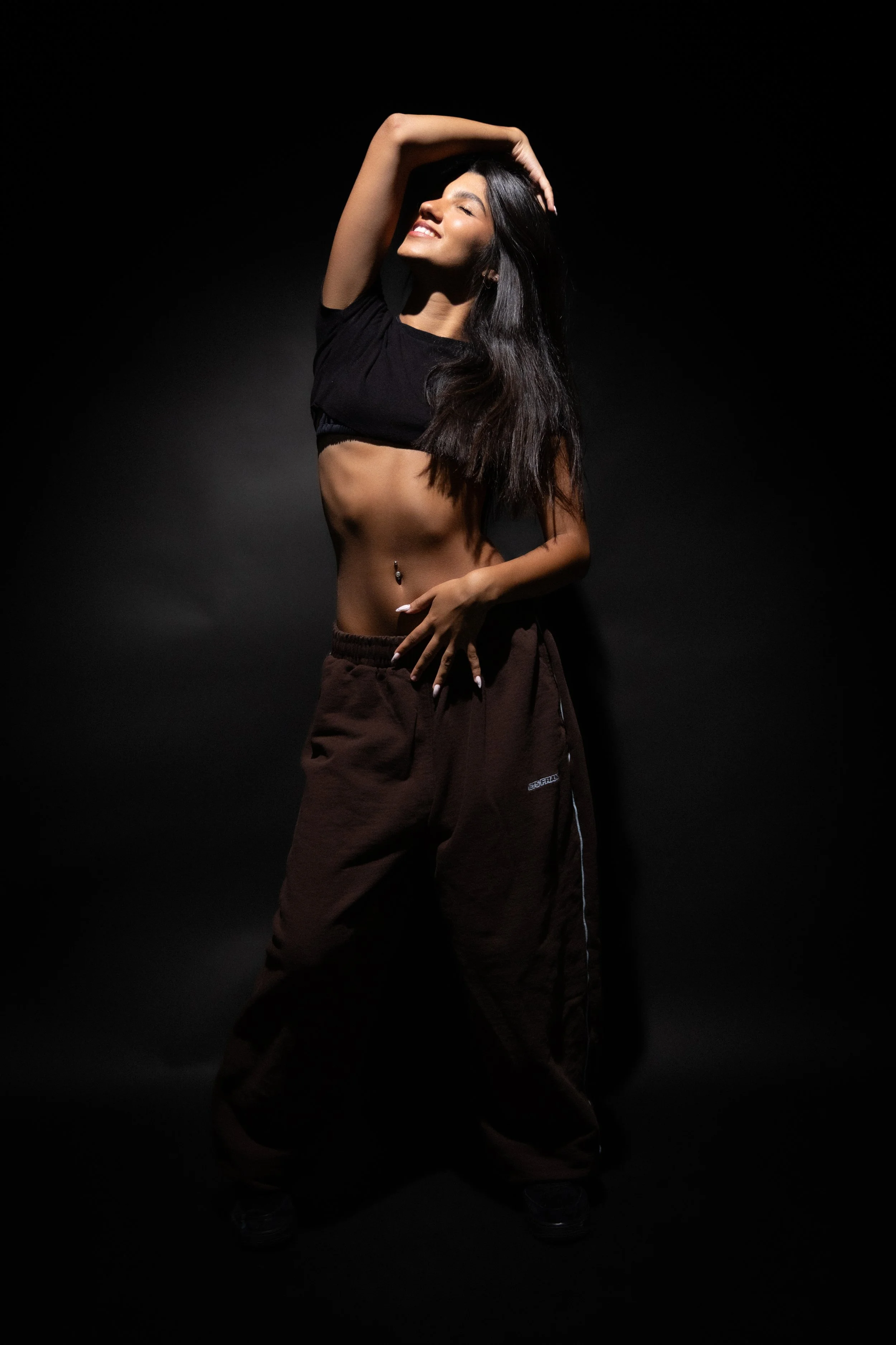 A woman with long dark hair is standing against a dark background, smiling with her eyes closed, wearing a cropped black t-shirt and brown loose-fitting pants, with one arm raised and hand on her head.