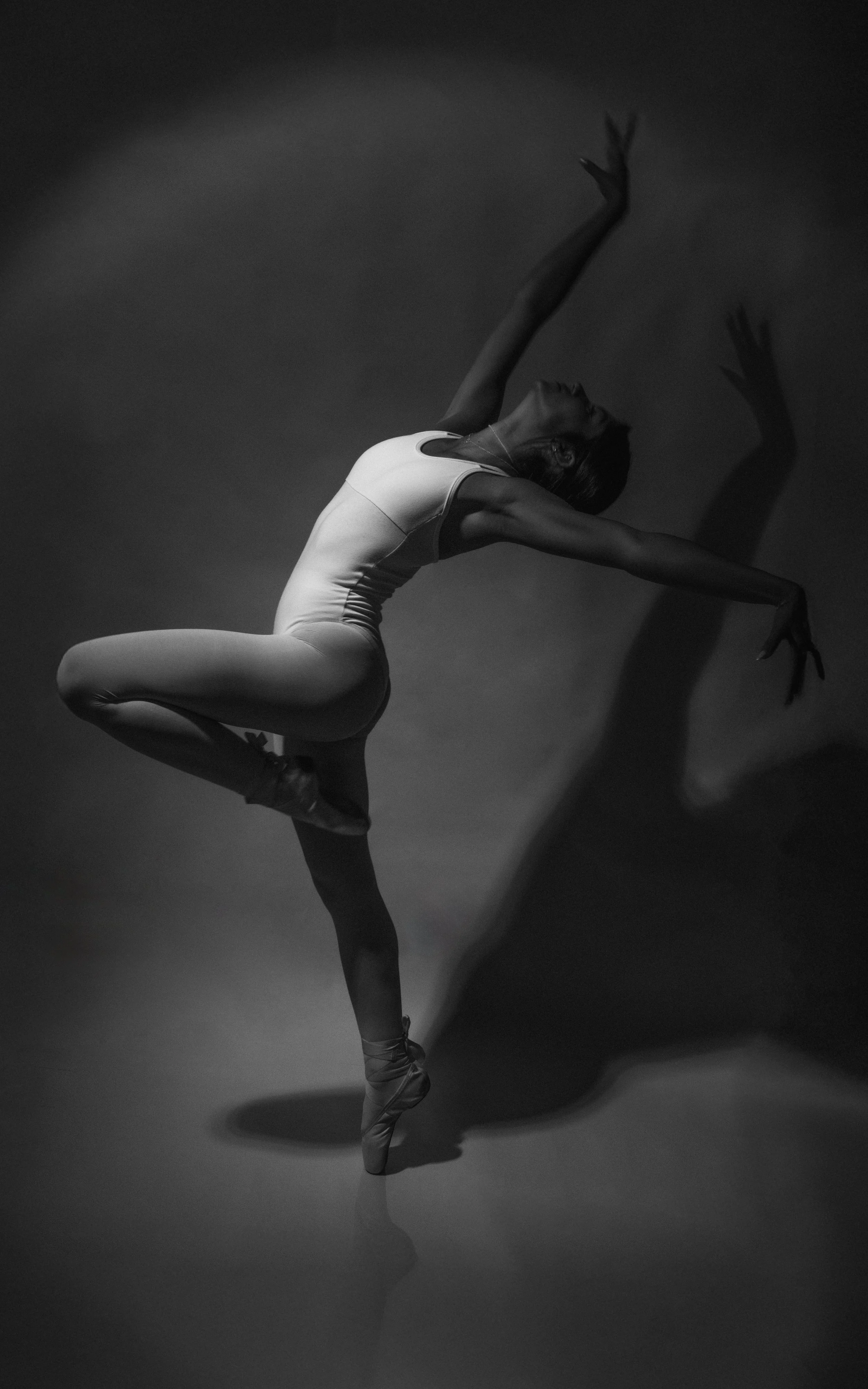 A ballet dancer practicing on stage, standing on one leg with arms extended, wearing a white leotard and pointe shoes, casting a shadow on the floor.