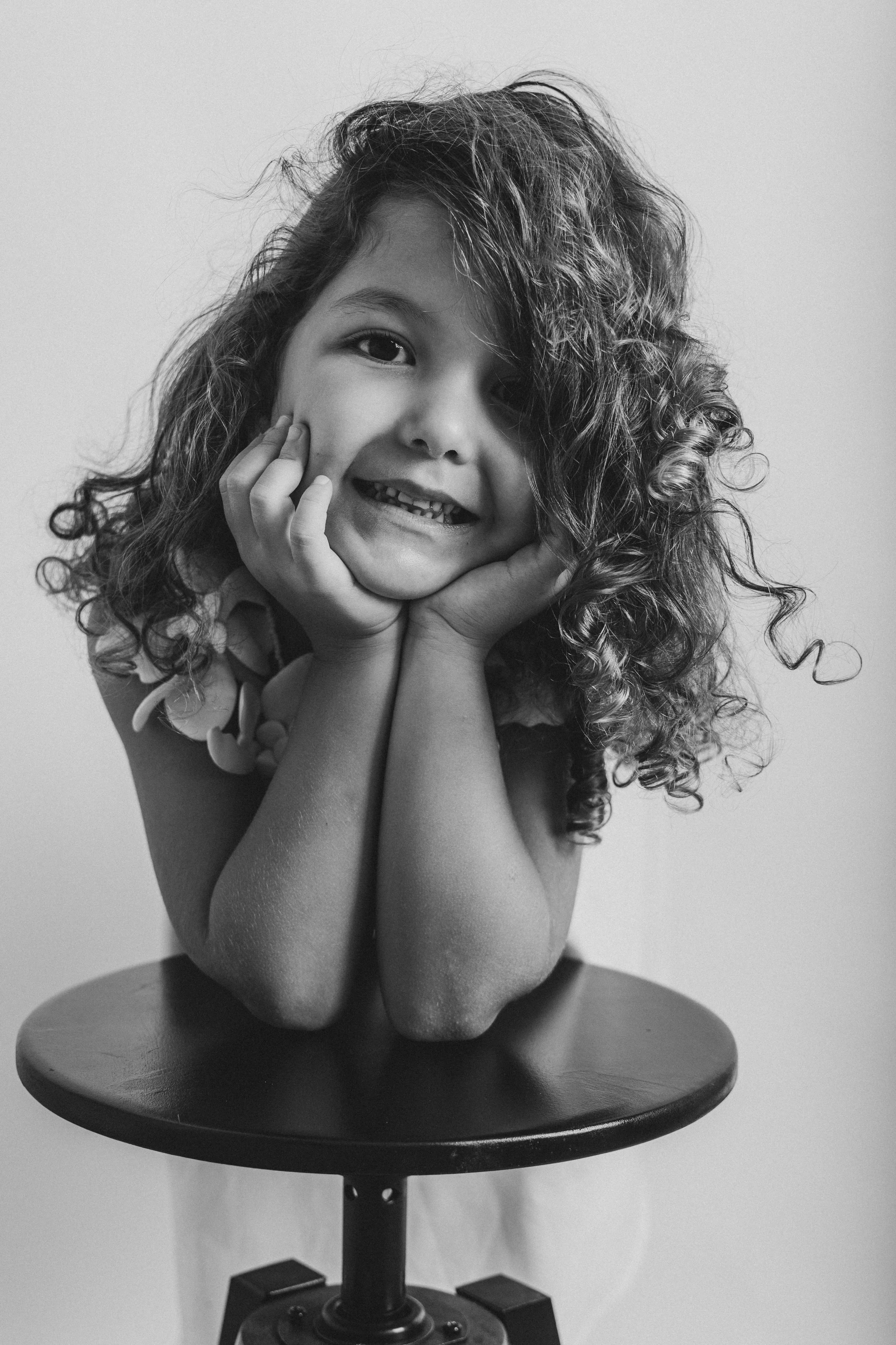 A monochrome sculpture of a smiling young girl with curly hair, resting her face in her hands, sitting on a round table with a plain background.