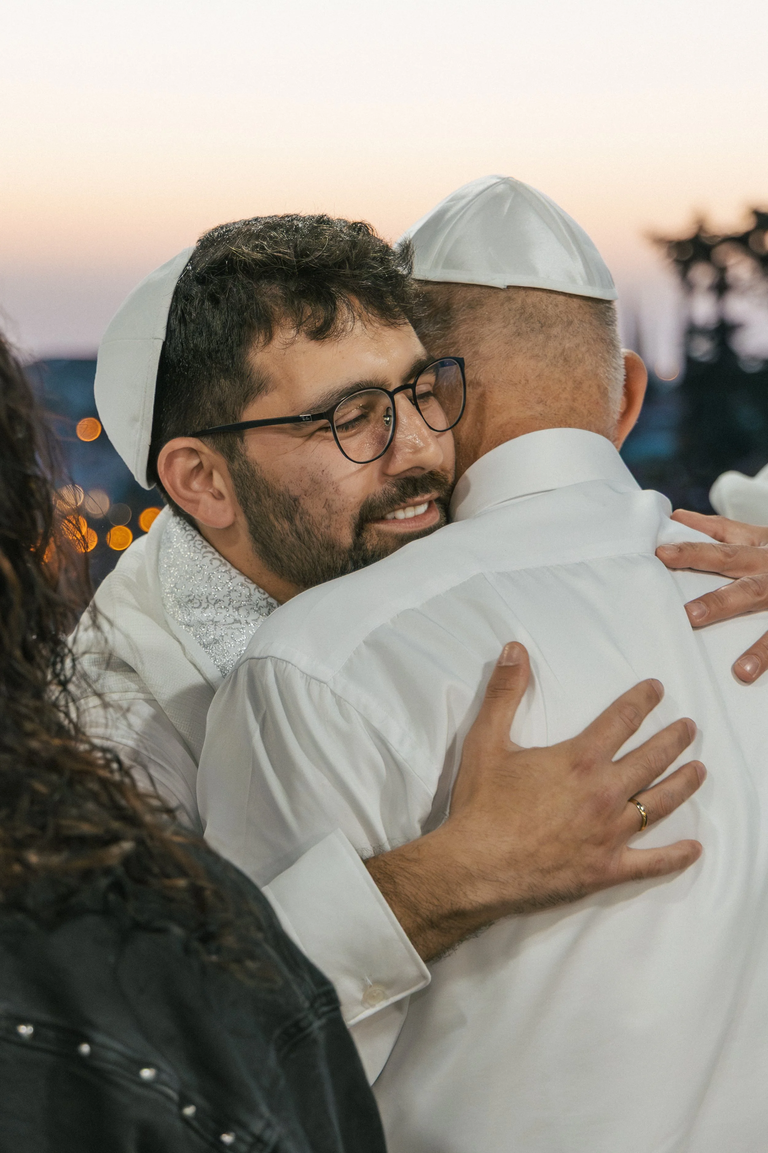 Two men embracing, one with glasses and a beard and the other with short hair and a yarmulke, outdoors during sunset.