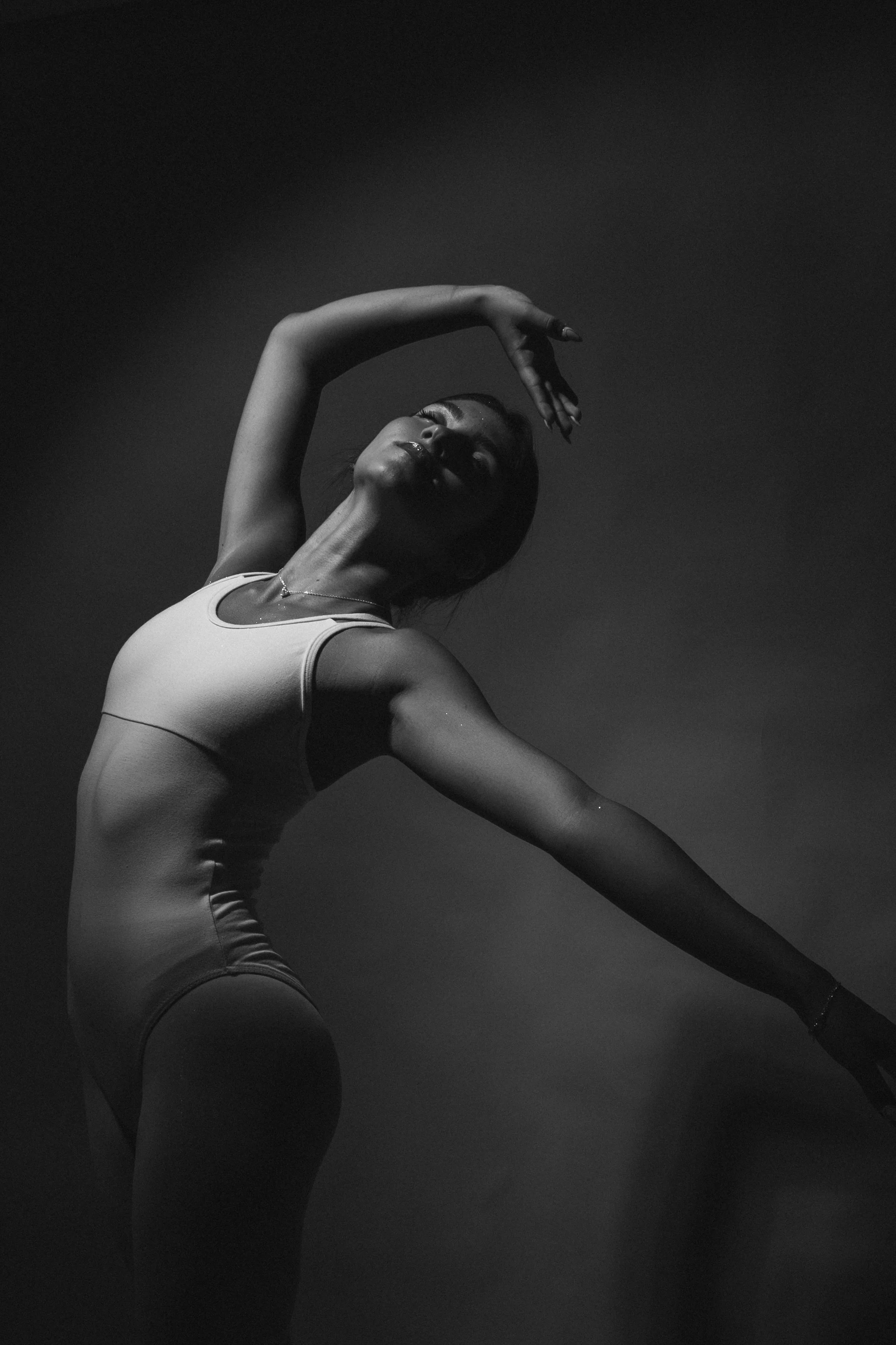 Black and white photo of a woman in athletic wear striking a dance pose with one arm above her head and her face tilted back.