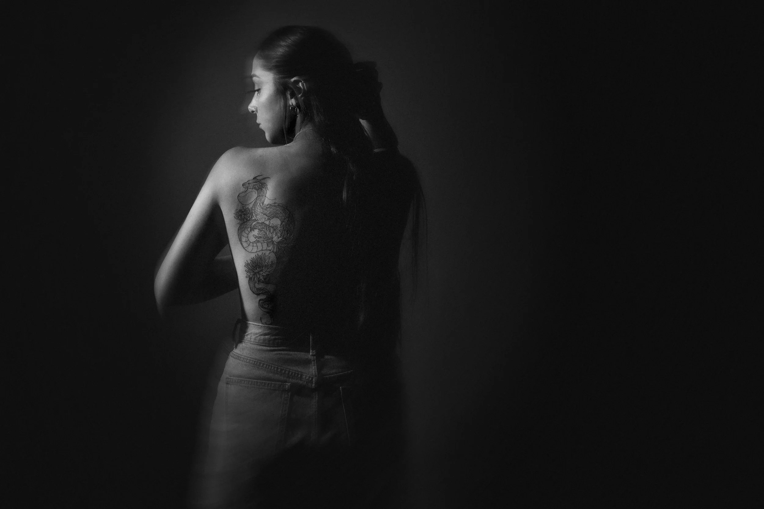 Black and white photo of a woman with long dark hair, standing sideways with her eyes closed, wearing a sleeveless top and jeans, with a dragon tattoo on her back, against a dark background.