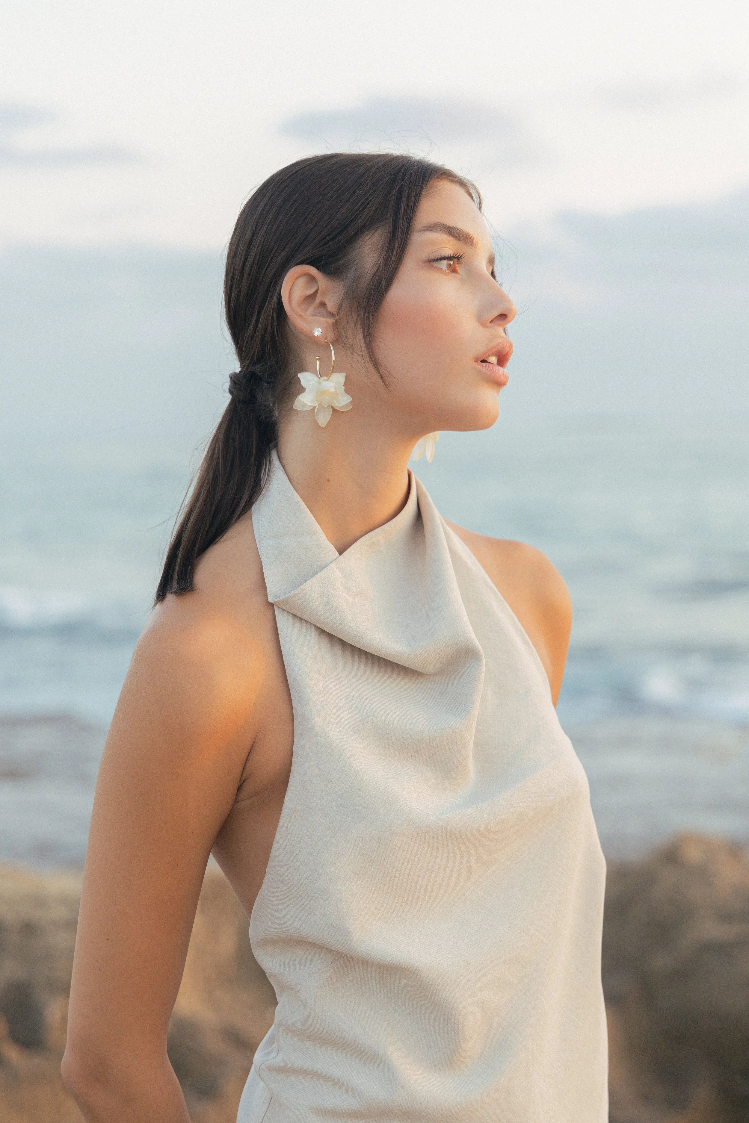 A young woman with long dark hair, wearing large flower-shaped earrings and a white sleeveless dress, looking to the right while standing on a beach with the ocean in the background.