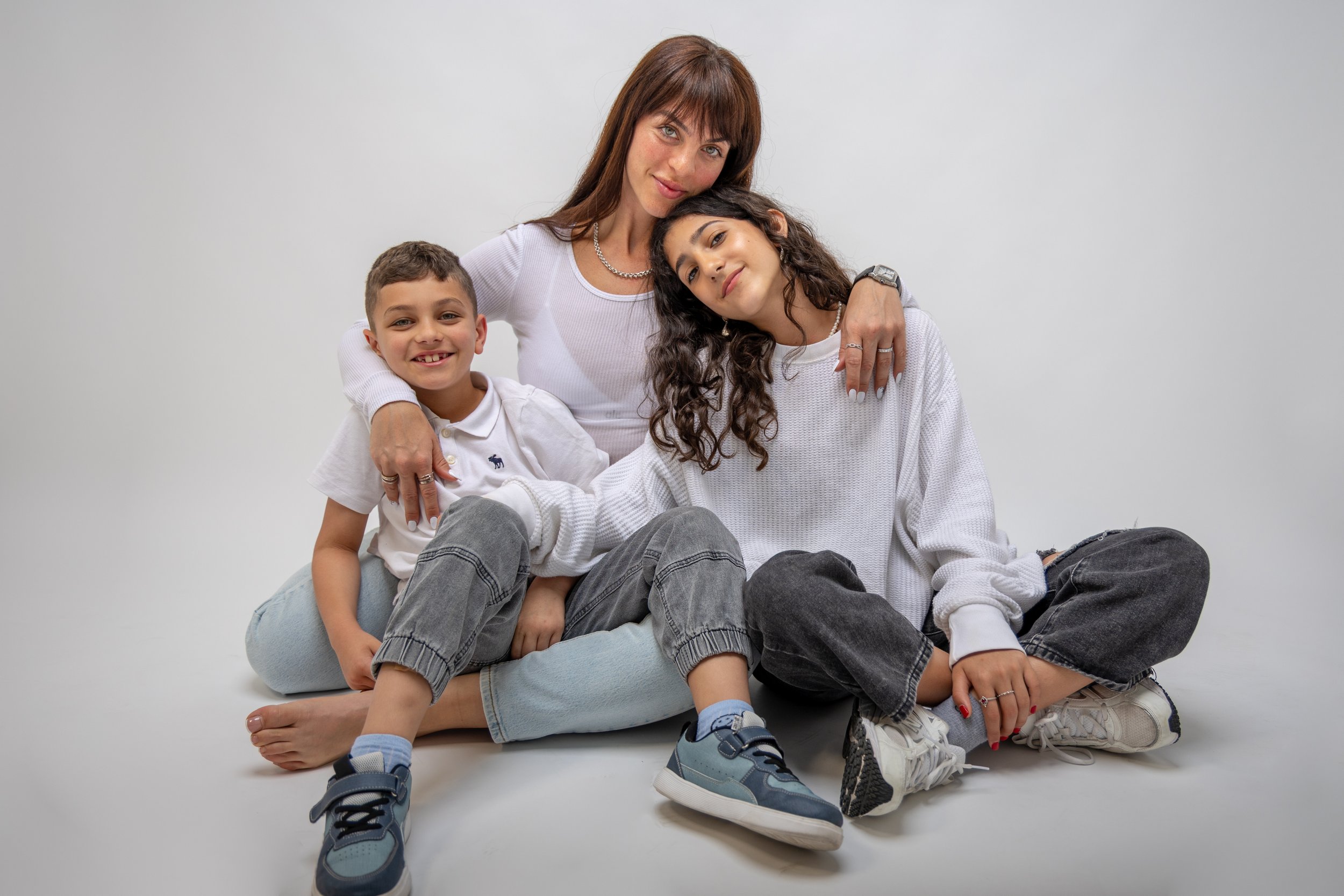 A woman with shoulder-length brown hair and a silver necklace sits on the floor with two children against a plain white background. The woman has her arm around a girl with curly dark hair, and a boy with short brown hair sits to the boy's left. They all wear casual white or light-colored clothing and are smiling or looking relaxed.
