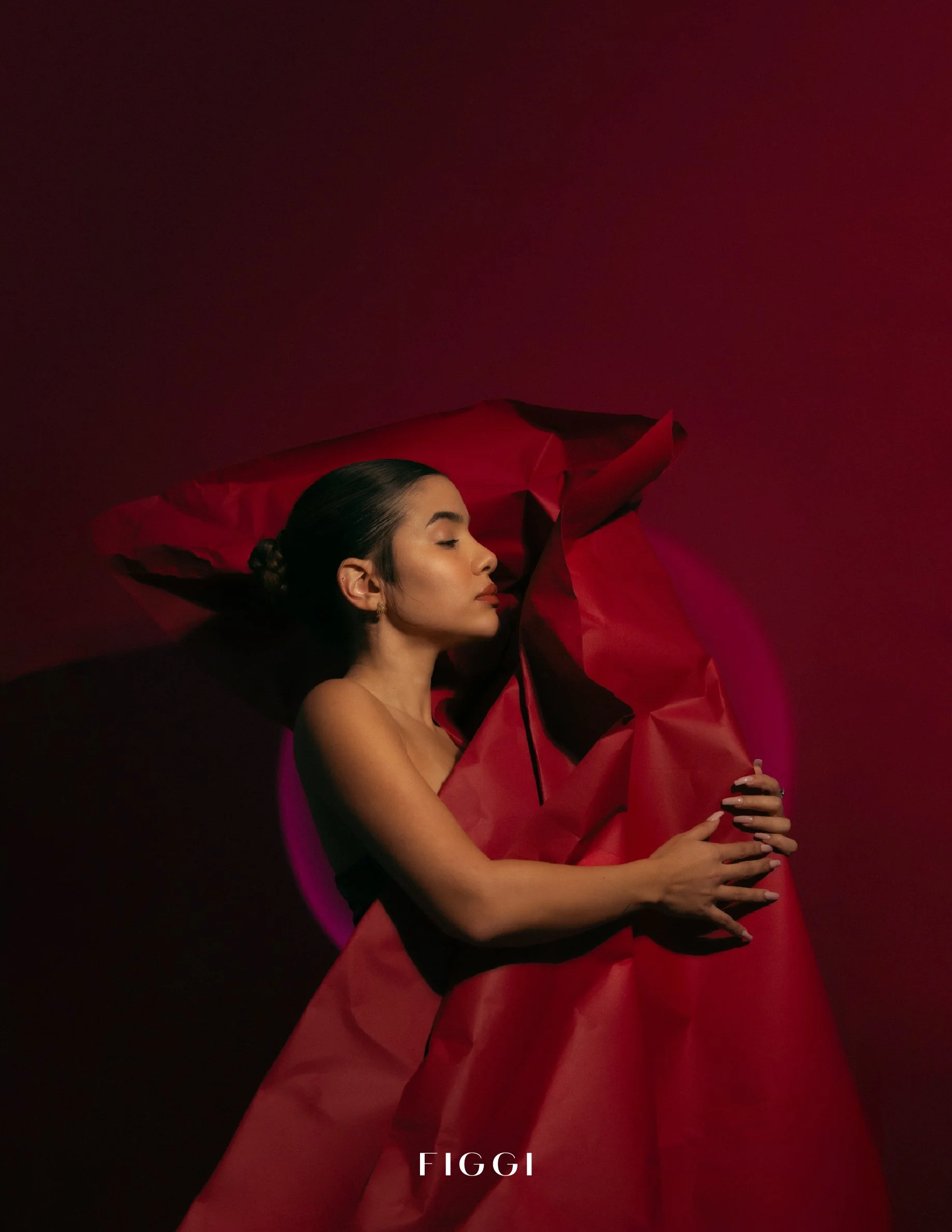 A woman with dark hair styled in a bun with eyes closed, holding a large, flowing red garment against a dark background with a red hue. The word 'FIGGI' is written at the bottom of the image.