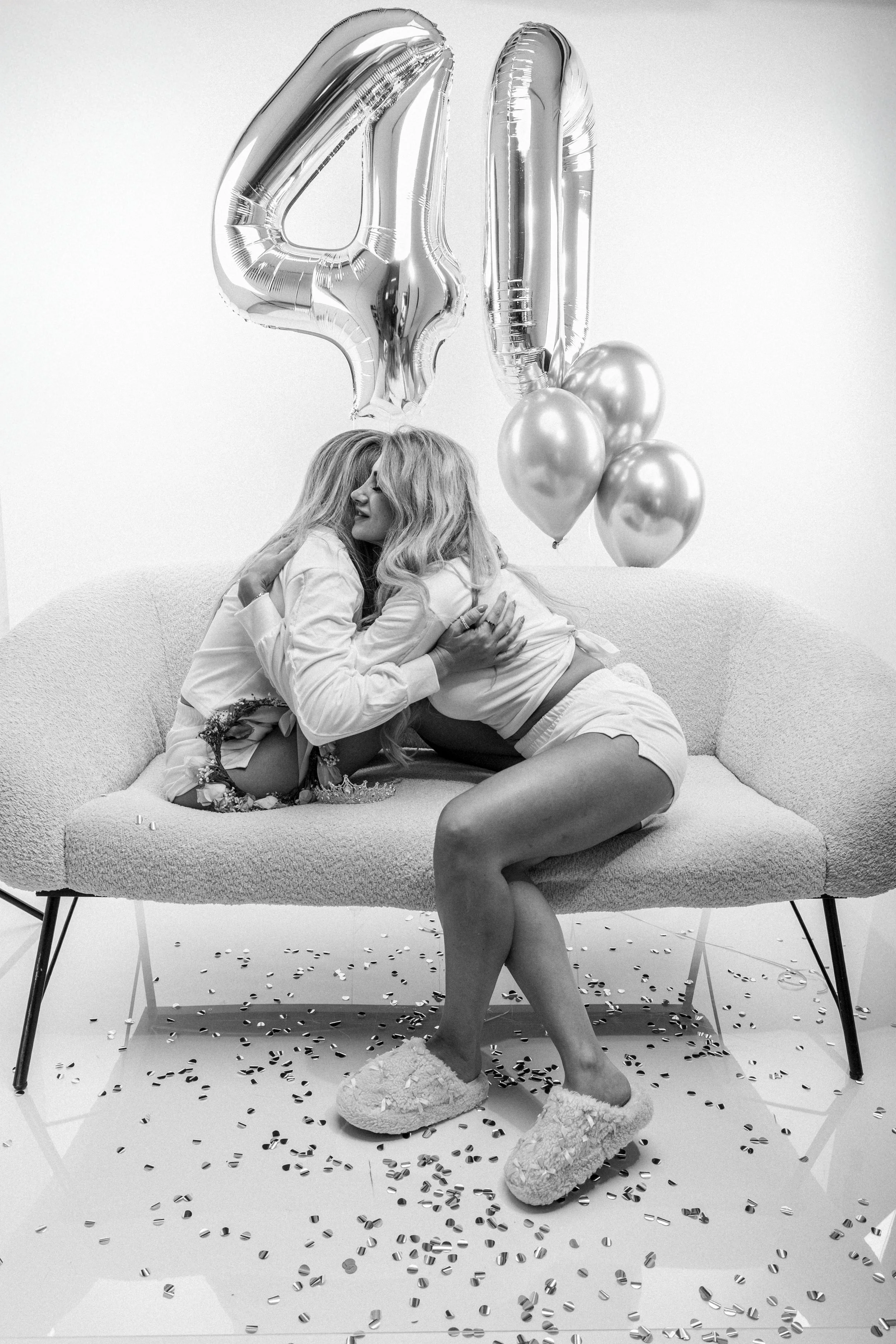 Two women sitting on a sofa, hugging each other, with balloons and confetti around them. One woman has long, blonde hair and the other has light-colored hair. The background is plain and decorated for celebration.