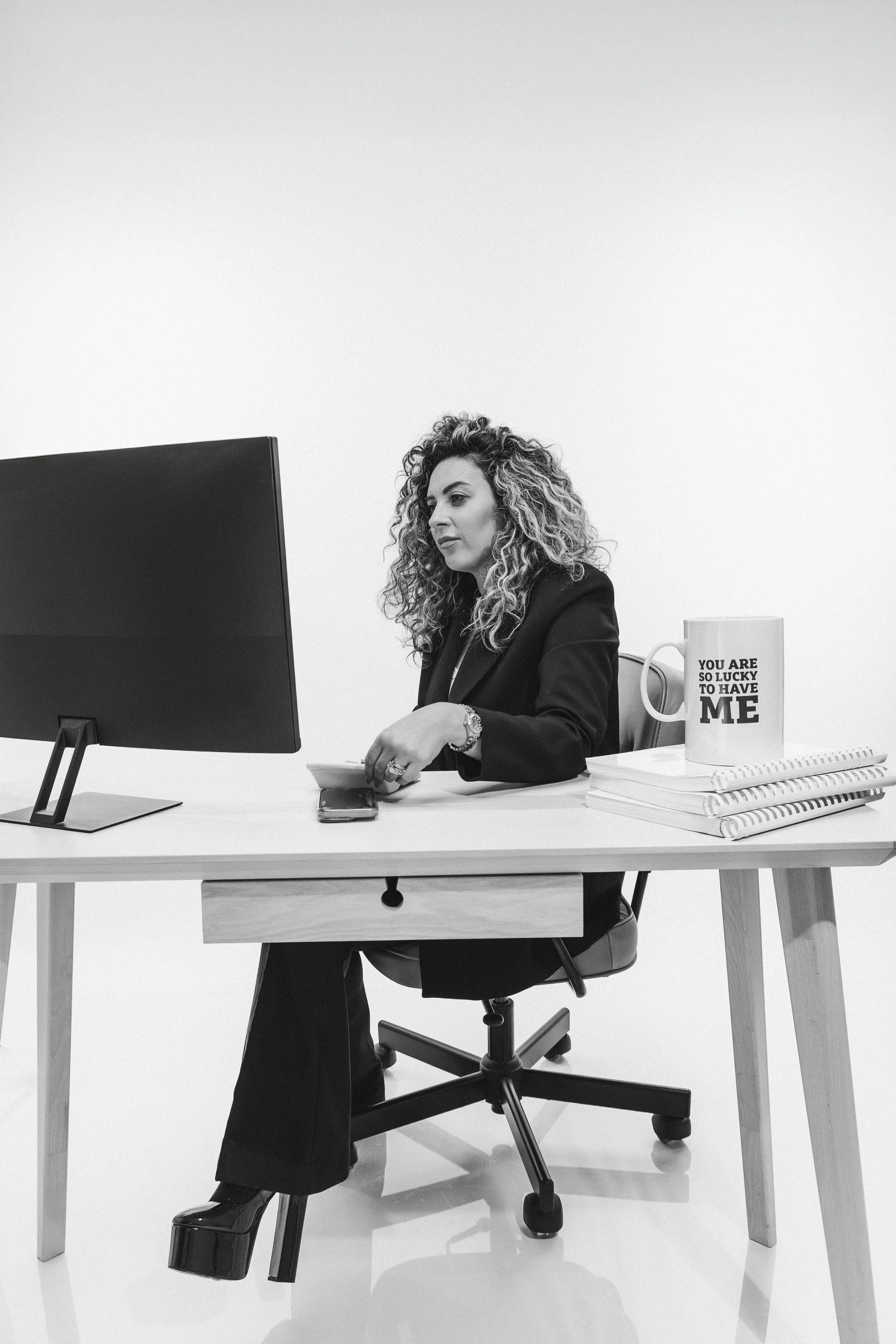 Black and white photo of a woman with curly hair working at a desk with a computer, mug, and notepad.
