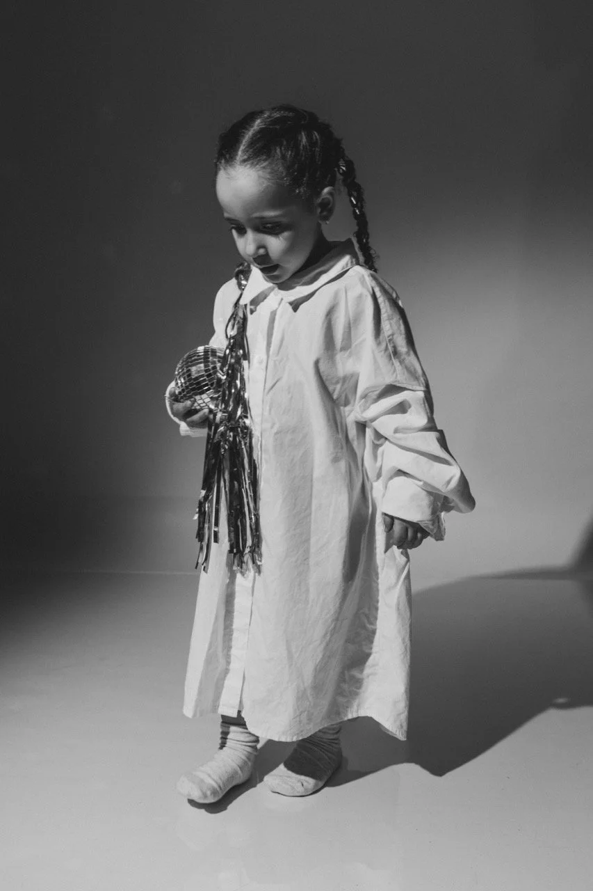 A young girl with braided hair, looking down and smiling, dressed in a long, light-colored dress, holding a small flag with tassels in her hand, standing on a plain surface.