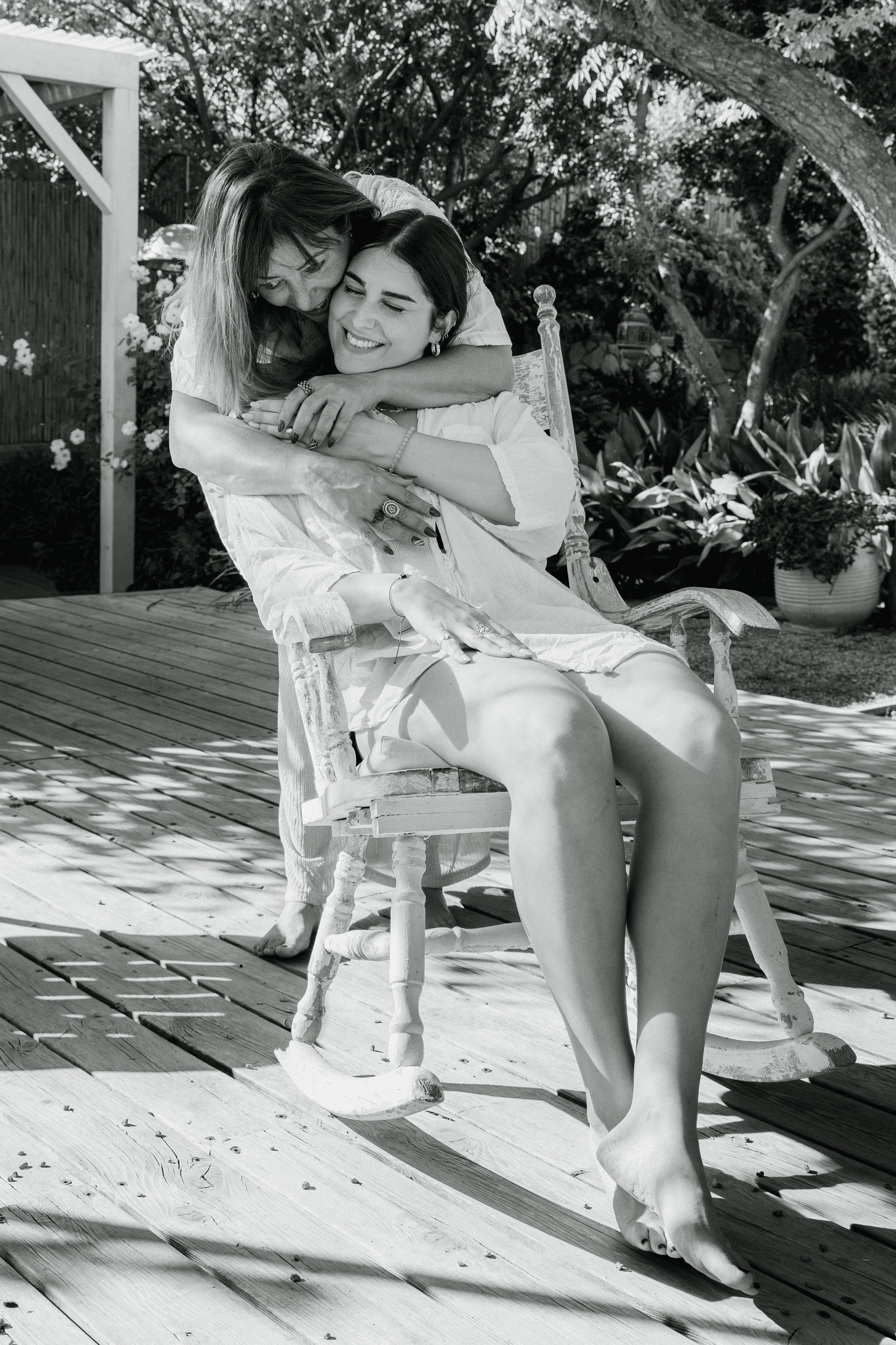 Two women sharing a joyful hug outdoors on a wooden deck, surrounded by plants and trees in a garden.
