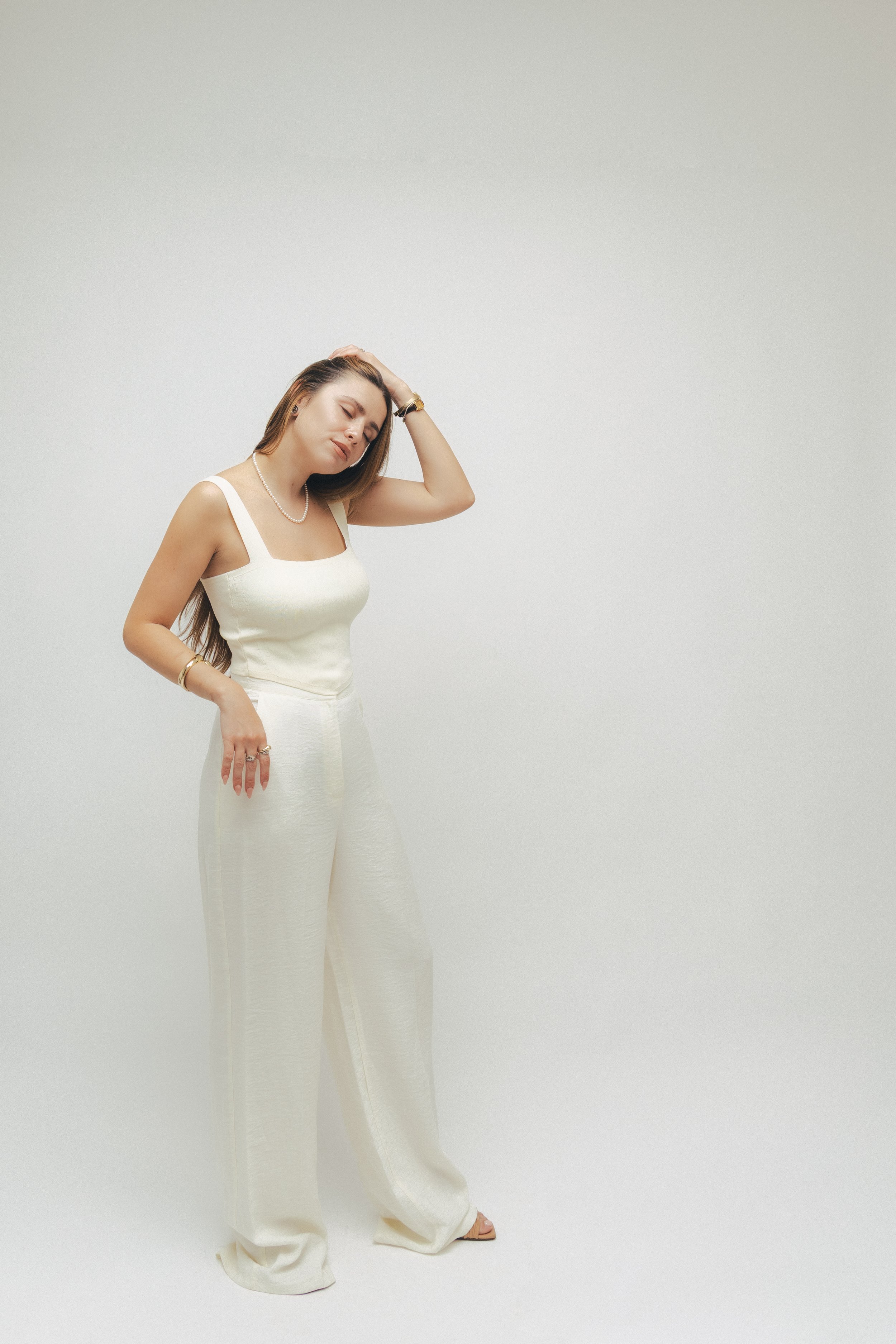 A woman dressed in an elegant white sleeveless top and wide-legged pants, wearing jewelry including a pearl necklace, bracelets, and rings, stands with her eyes closed and one hand on her head against a plain white background.