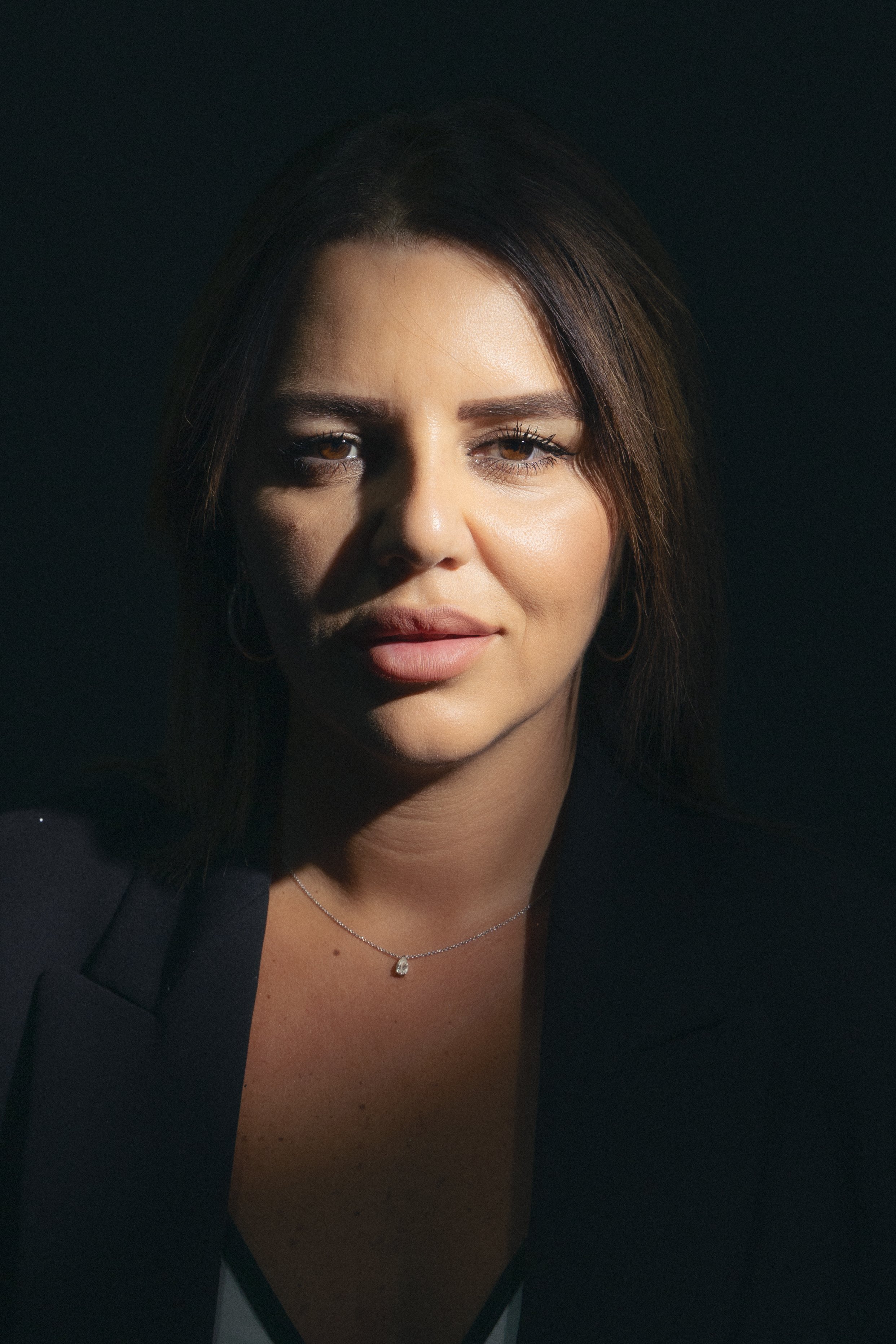 Close-up of a woman with dark hair, wearing makeup, a black blazer, a necklace with a teardrop-shaped pendant, and a black top or dress. She has a slight smile and is looking directly at the camera against a dark background.
