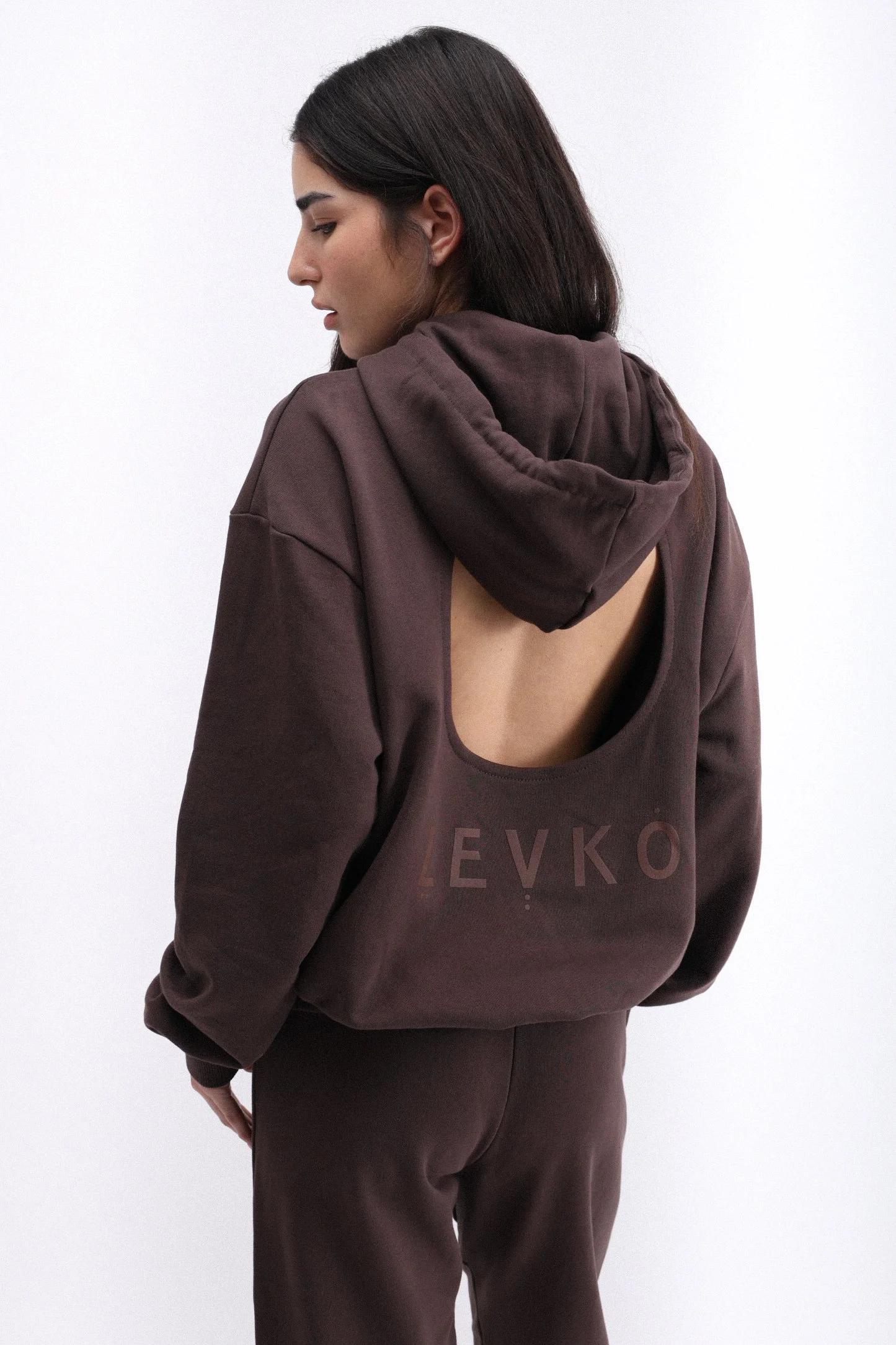 A woman with dark hair wearing a brown hoodie with a large open back and matching sweatpants, standing against a plain white background.