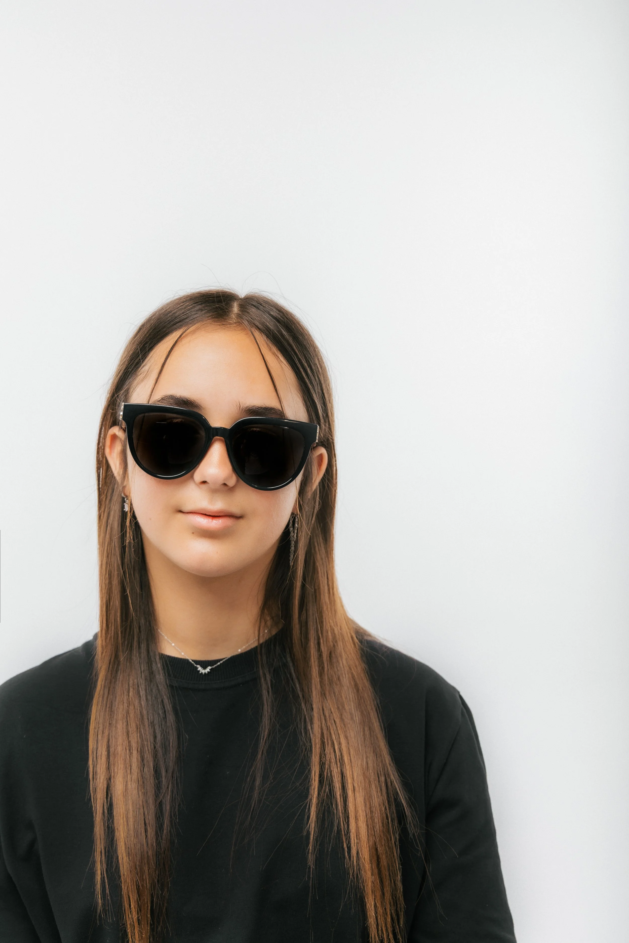 A young woman with long brown hair wearing black sunglasses, a black shirt, and a delicate necklace, standing against a plain white background.