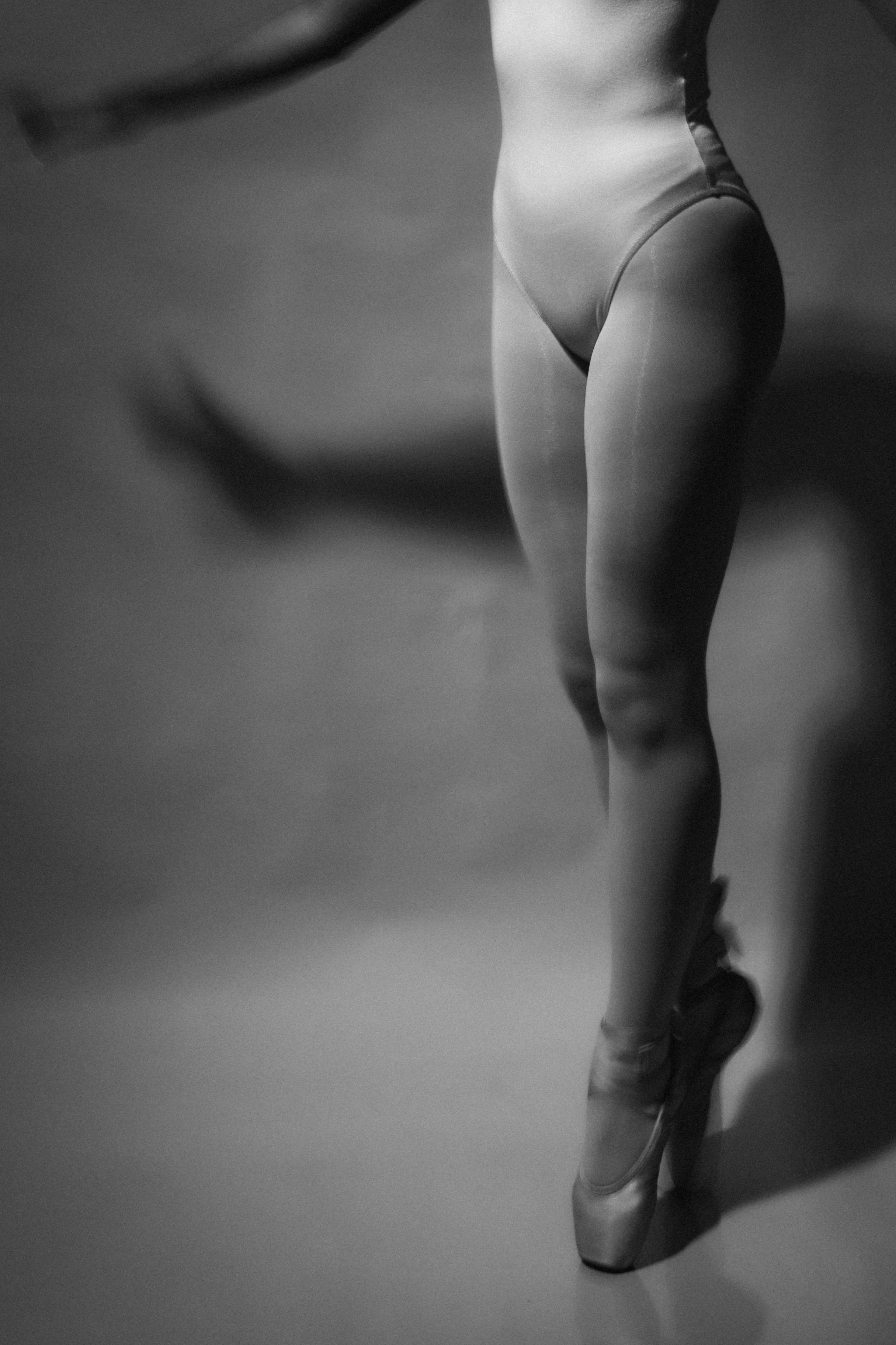 Black and white photo of a ballet dancer's lower body, wearing pointe shoes and a leotard, standing on her toes with one foot in front of the other against a plain background.