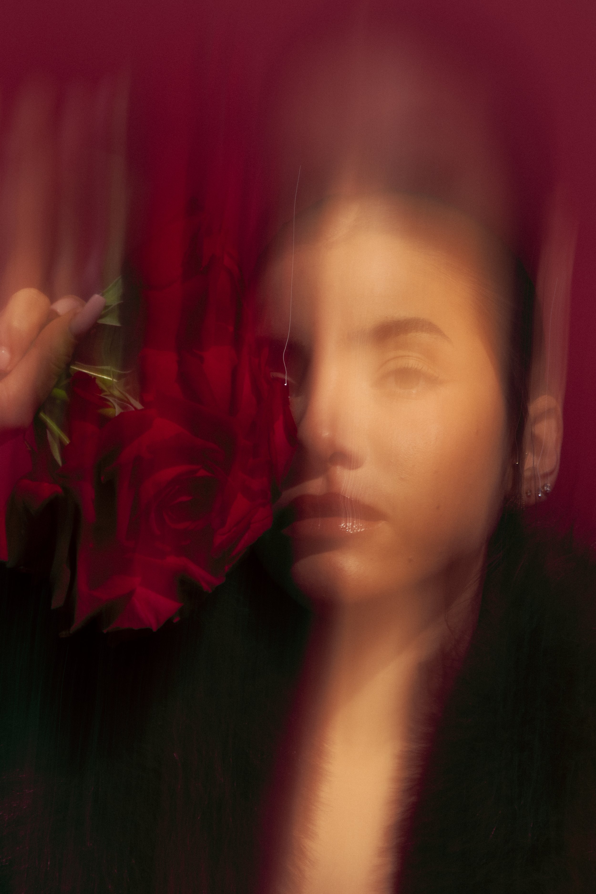 A woman with dark hair, partially covered by a red curtain and holding red roses, with soft makeup and earrings, in a dimly lit setting.