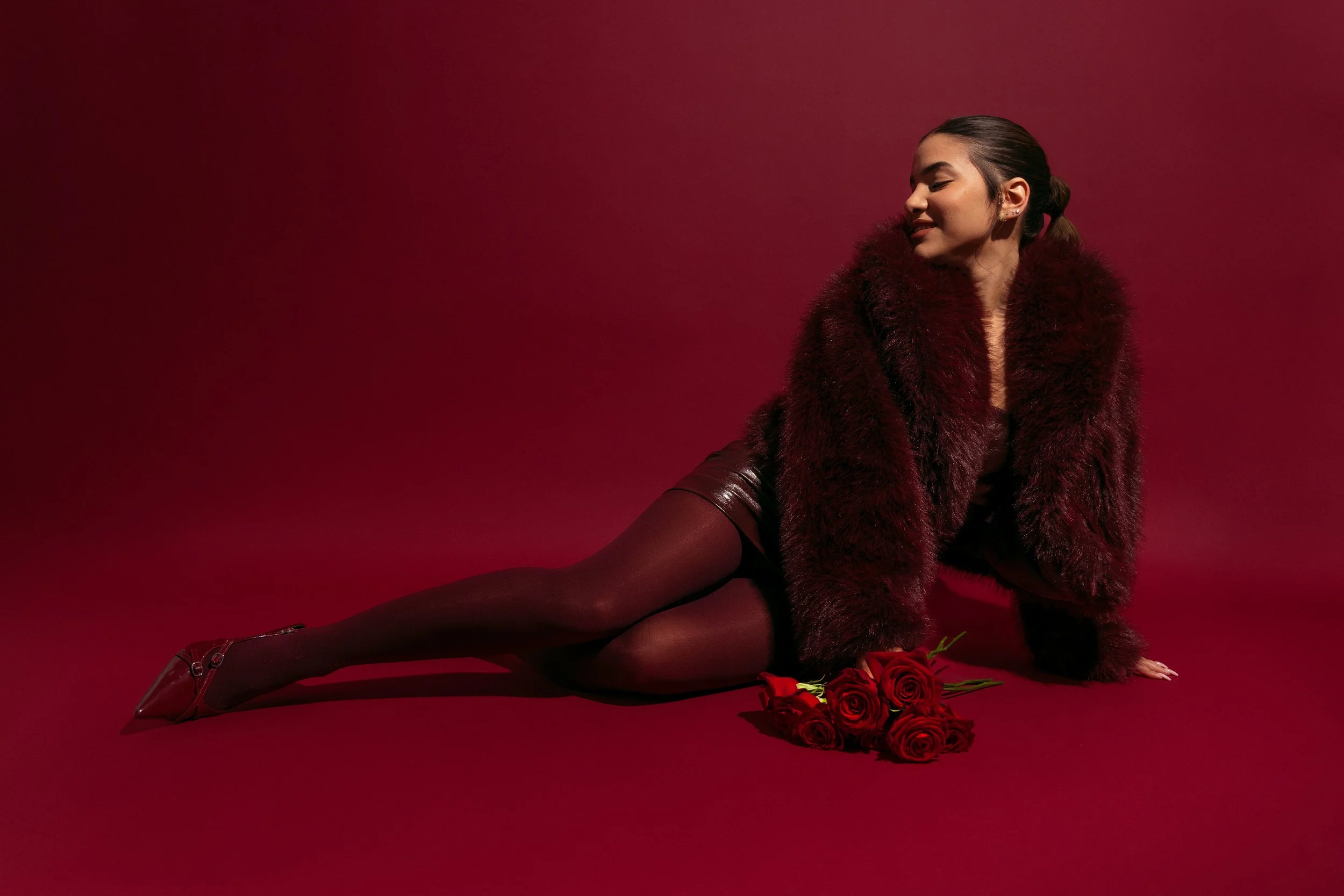 A woman with dark hair styled in a ponytail is sitting against a red background. She is wearing a dark red fur coat and black stockings, and is lying on her side with her eyes closed and a slight smile. There are red roses placed on the floor next to her.
