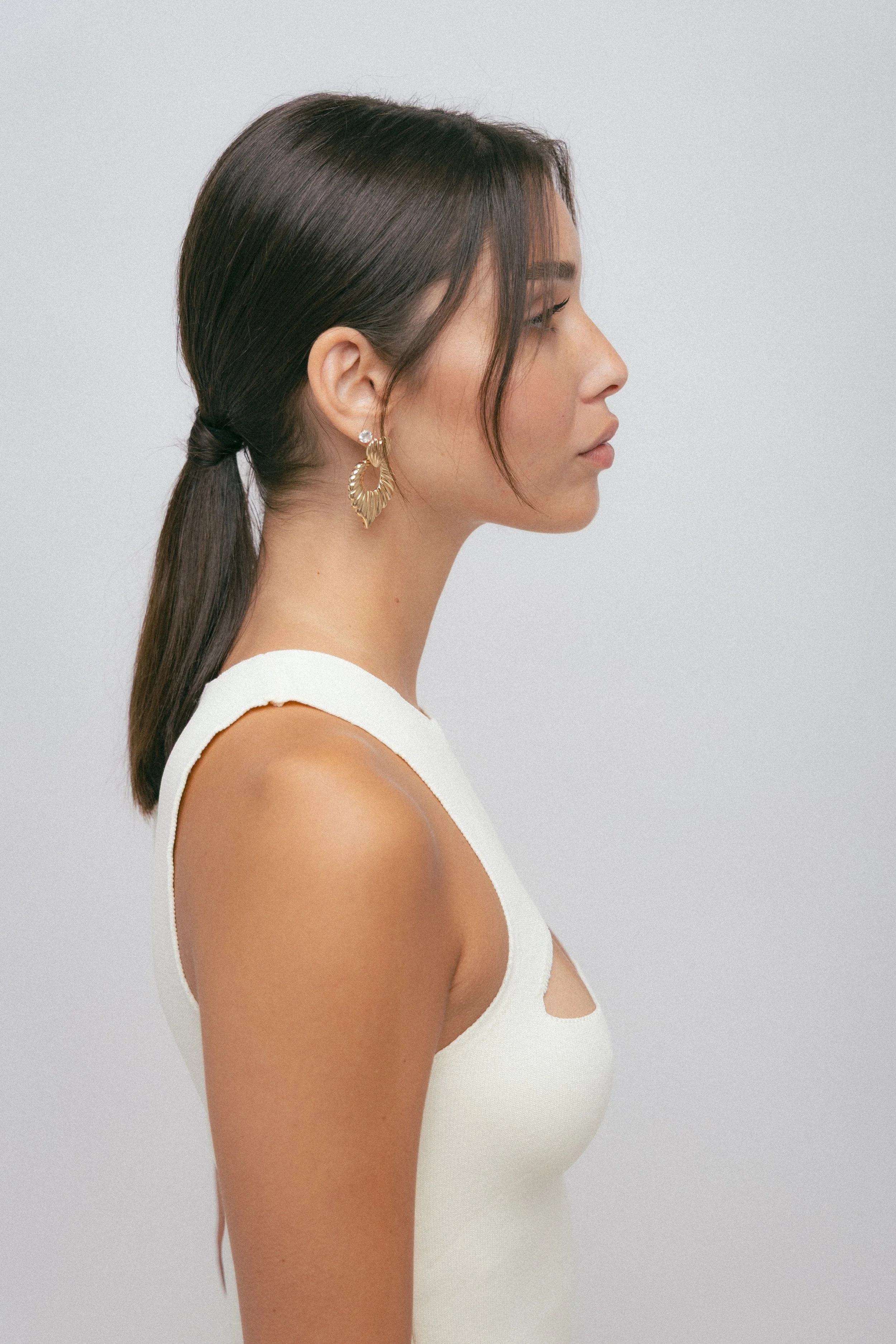 Profile of a woman with long dark hair tied back, wearing gold earrings and a white sleeveless top, standing against a plain light gray background.