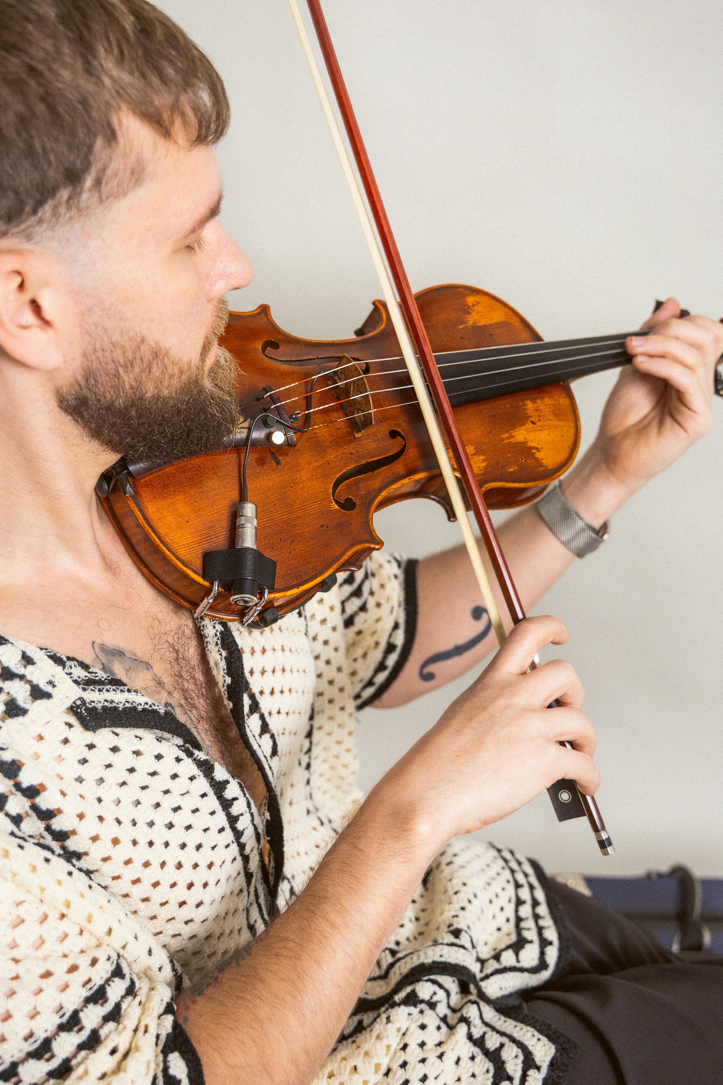 A man with a beard and tattoos playing a violin, wearing a patterned sweater.