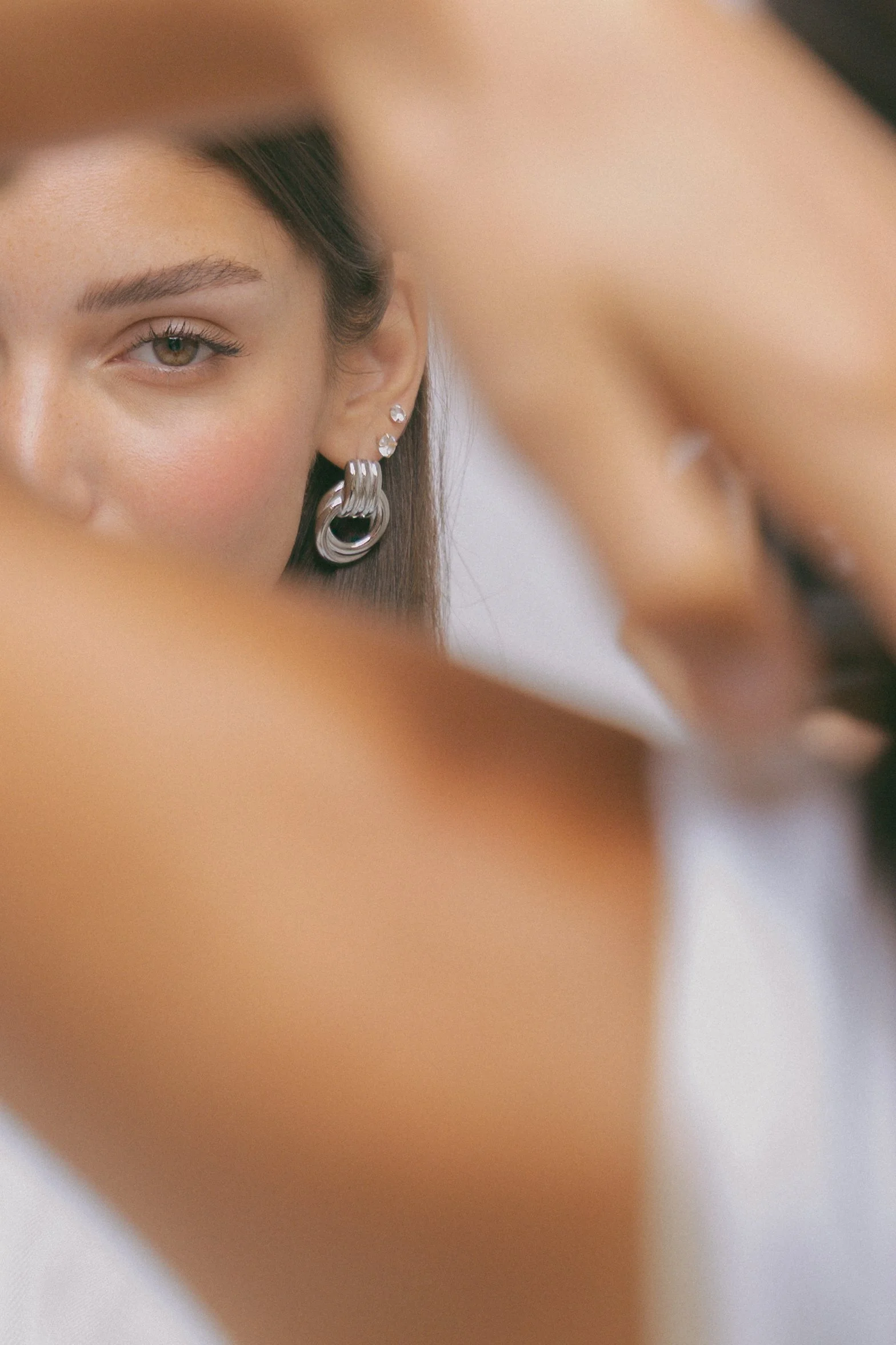 Close-up of a woman's face with one eye visible, wearing multiple earrings, and her hand partially obscuring the image.