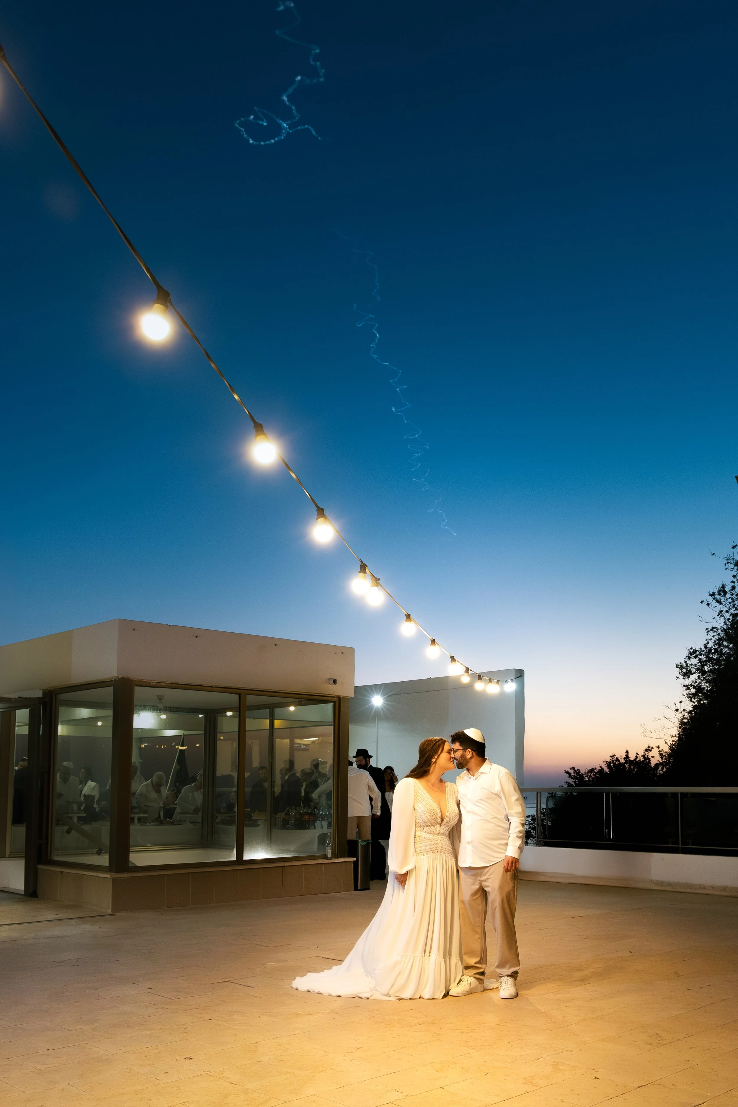 A couple dressed in elegant wedding attire stands close together, smiling and sharing a tender moment on a rooftop at sunset, with string lights above and a view of the evening sky with a faint cityscape in the background.