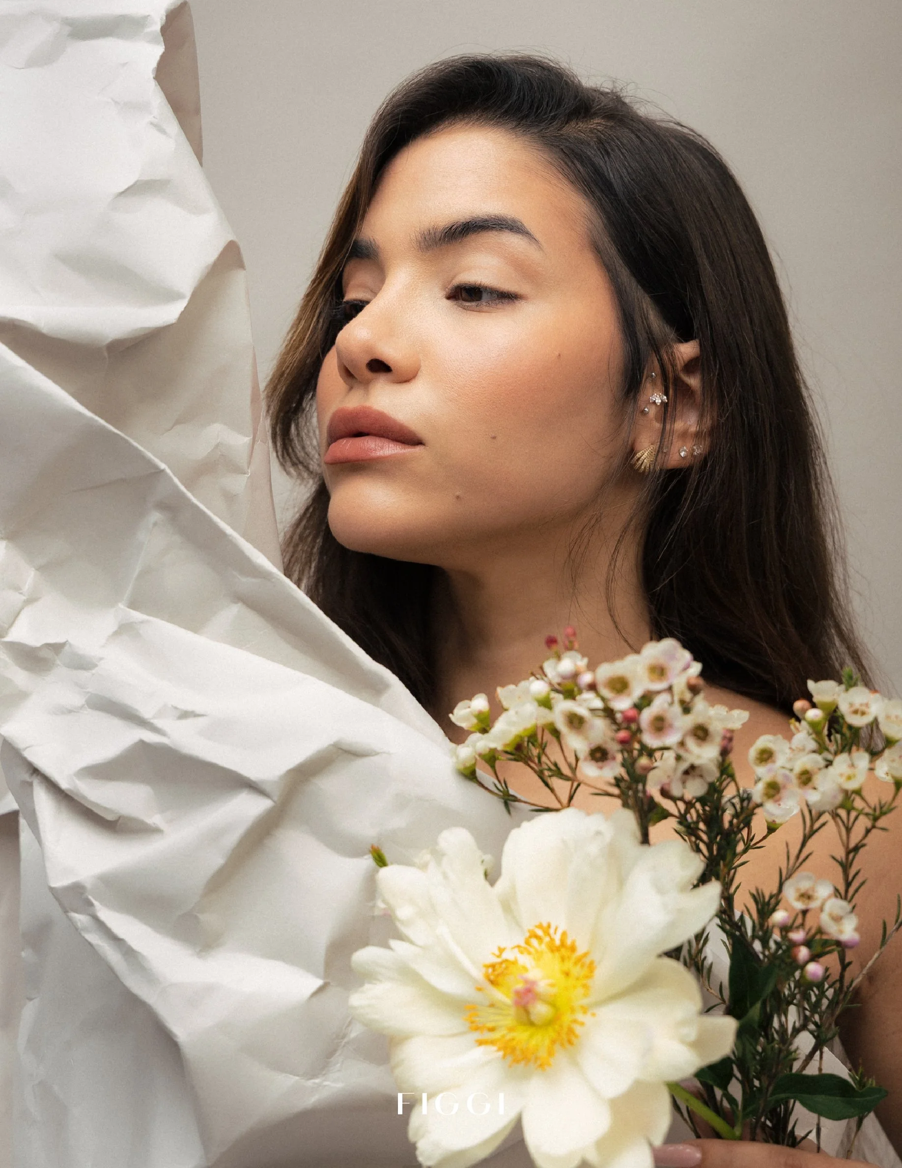 A woman with dark hair and earrings looking at a bouquet of white flowers with yellow centers, partially wrapped in crumpled white paper.