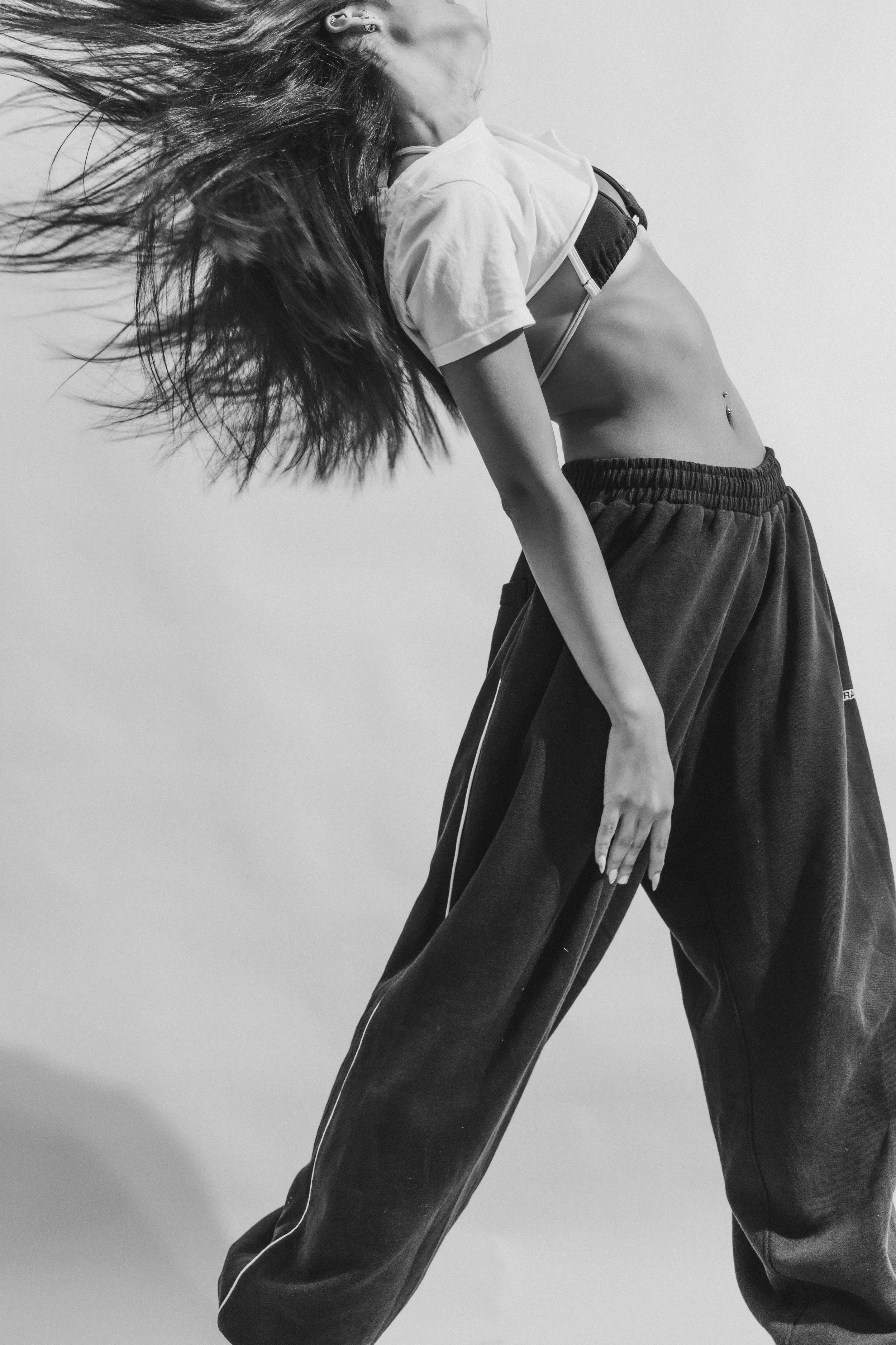 Black and white image of a woman with long hair moving, wearing a cropped t-shirt and baggy pants, standing with a hand on her hip against a plain background.