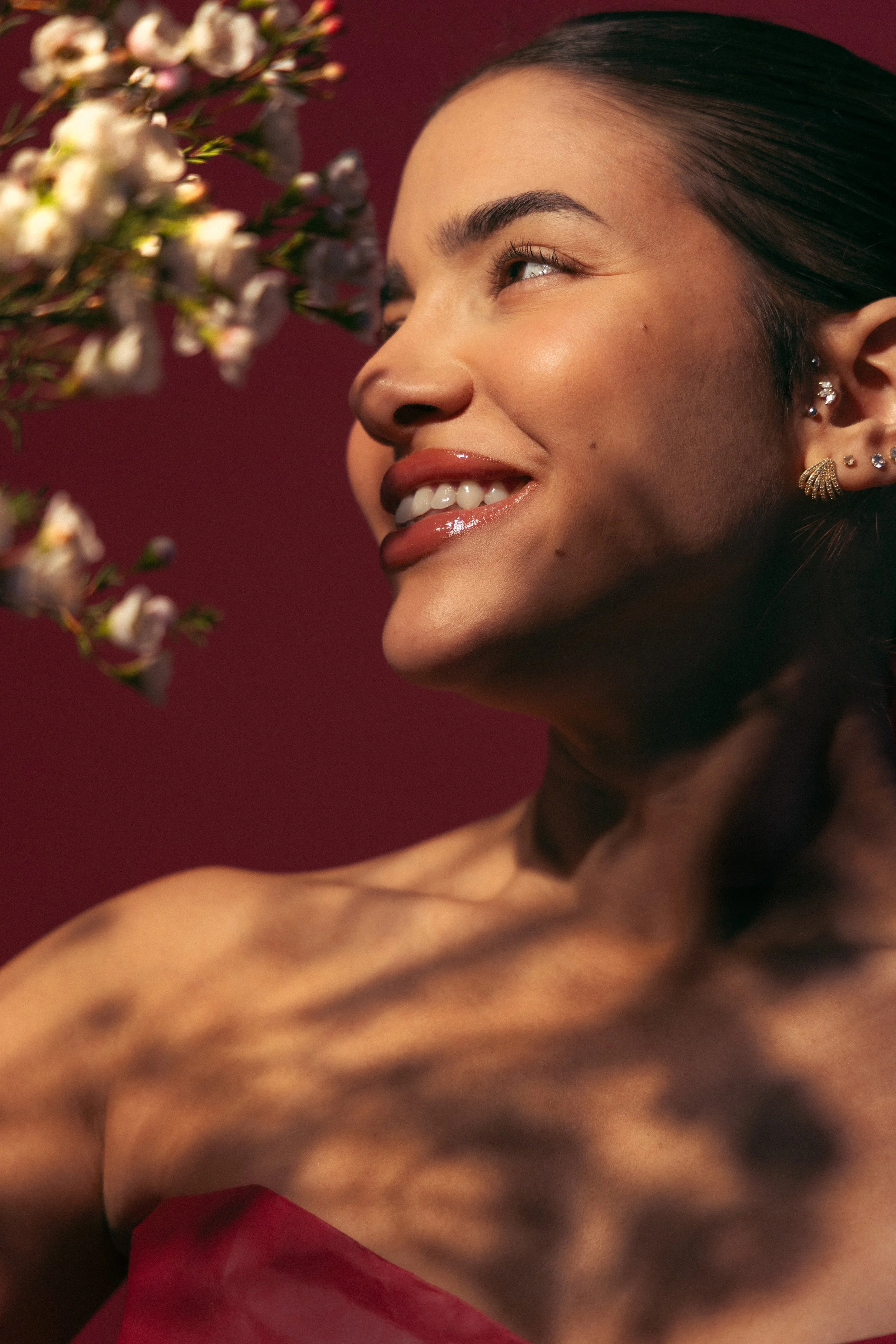 Close-up of a smiling woman with dark hair, multiple earrings, and smooth skin. She is looking slightly to the side with a blurred branch of flowers in the foreground. The background is a solid maroon color.