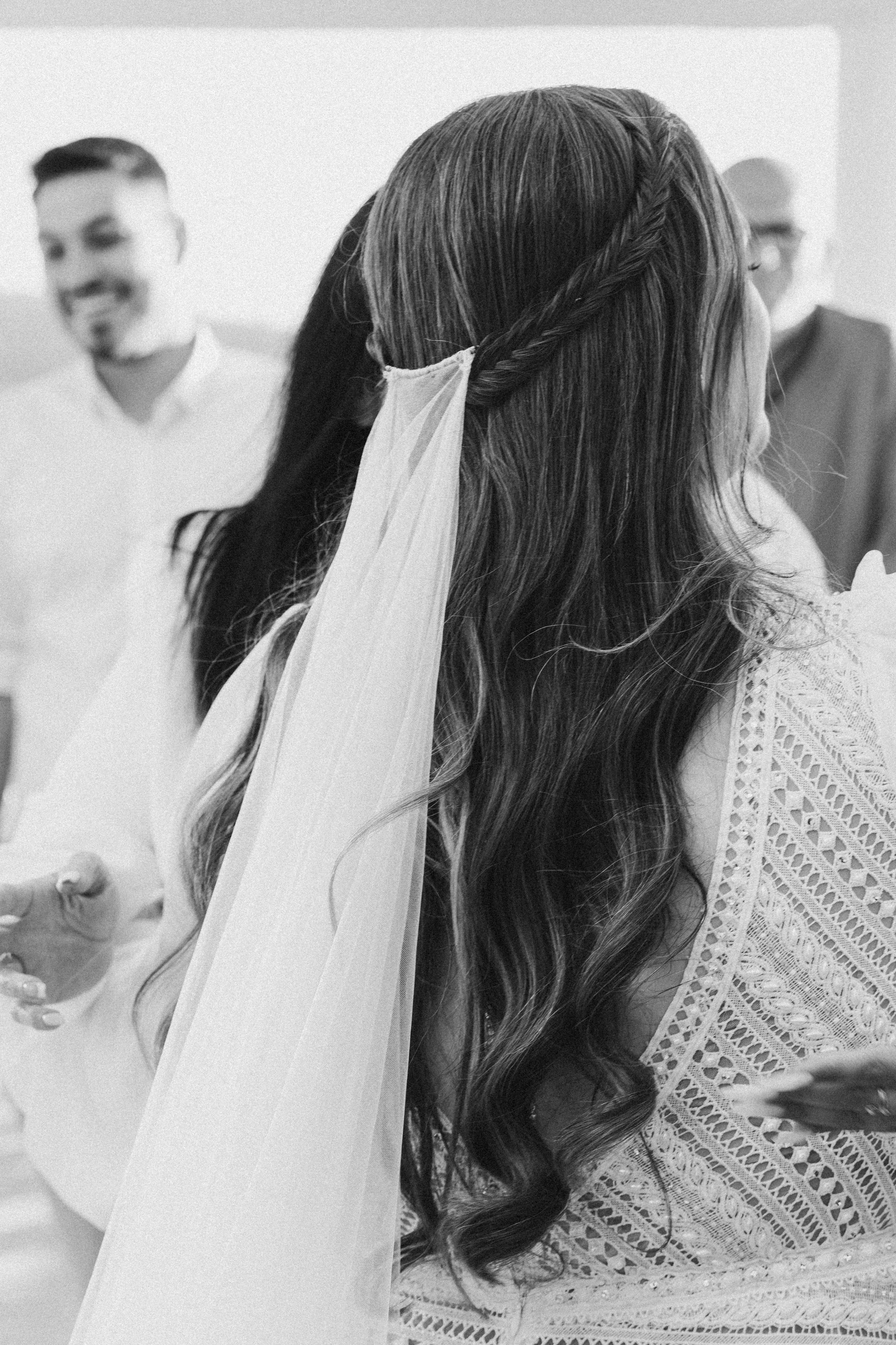 A woman with long curly hair in a braid and lace dress turns to the right during a wedding ceremony, while a man in a suit and another man in a white shirt smile in the background.