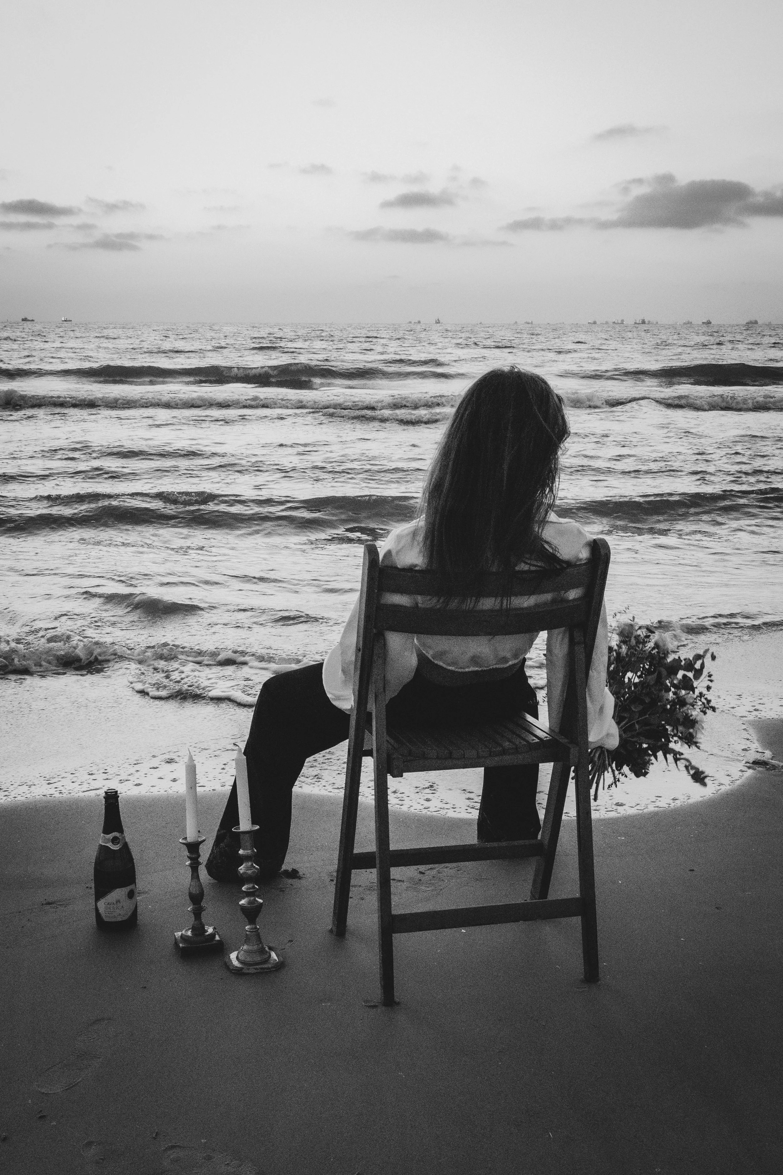 Person sitting on a chair facing the ocean, with candles and bottles on the ground nearby, during sunset or overcast day. artistic edit. model. outdoor photoshoot. צילומים בים, צילומי אופנה, צילום למגזין, דוגמנית, צילומי חוץ