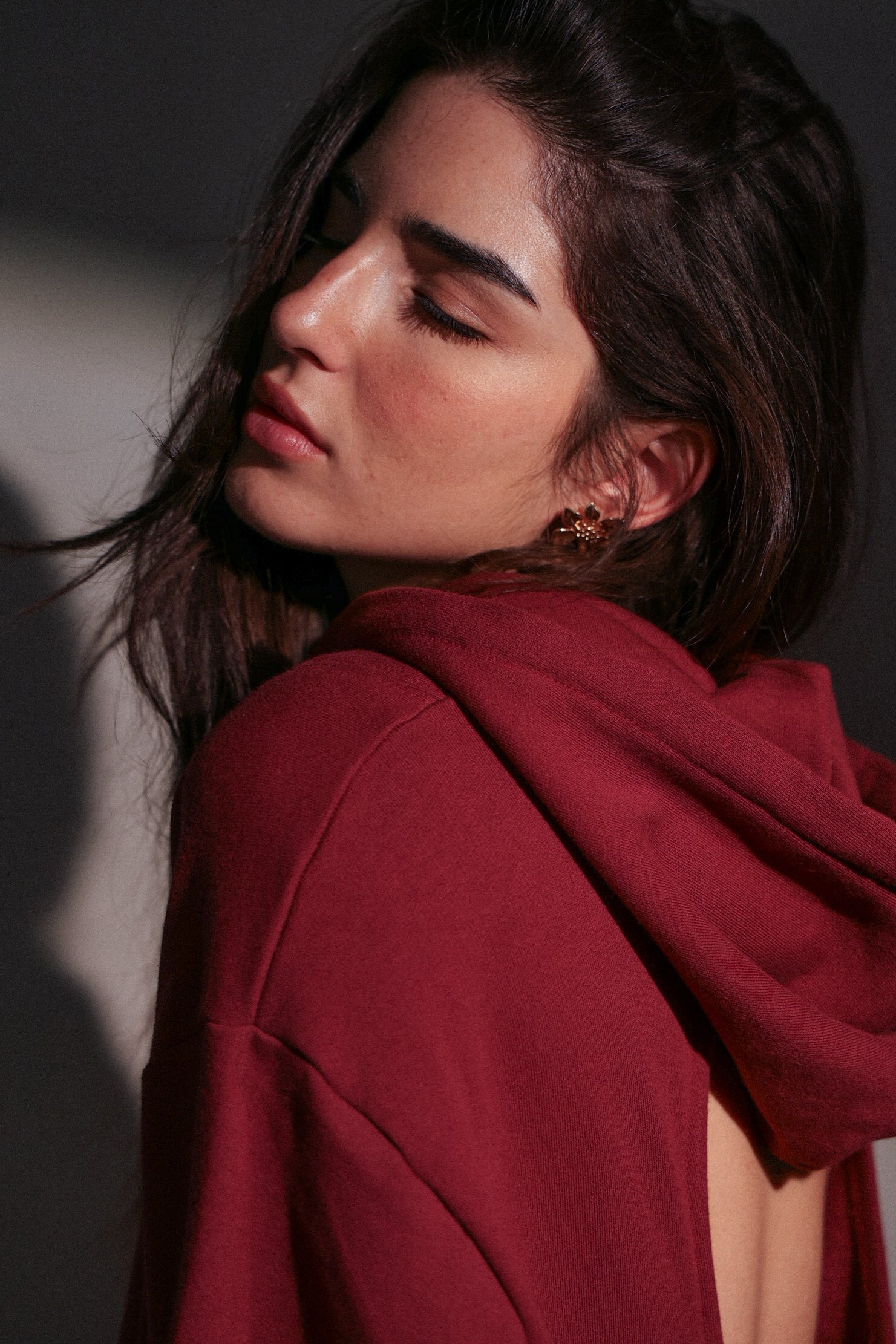 Close-up of a woman with long dark hair, closed eyes, and wearing gold earrings and a red hoodie.