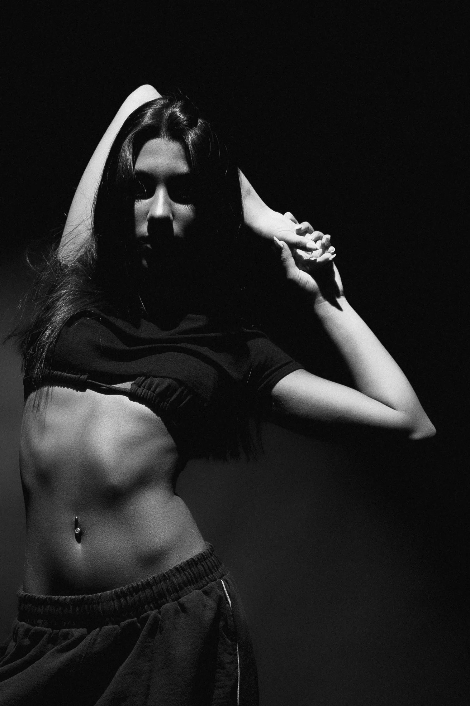 Black and white photo of a woman with long hair, holding her right hand near her face, wearing a sports bra and sweatpants, with her face partially shadowed.
