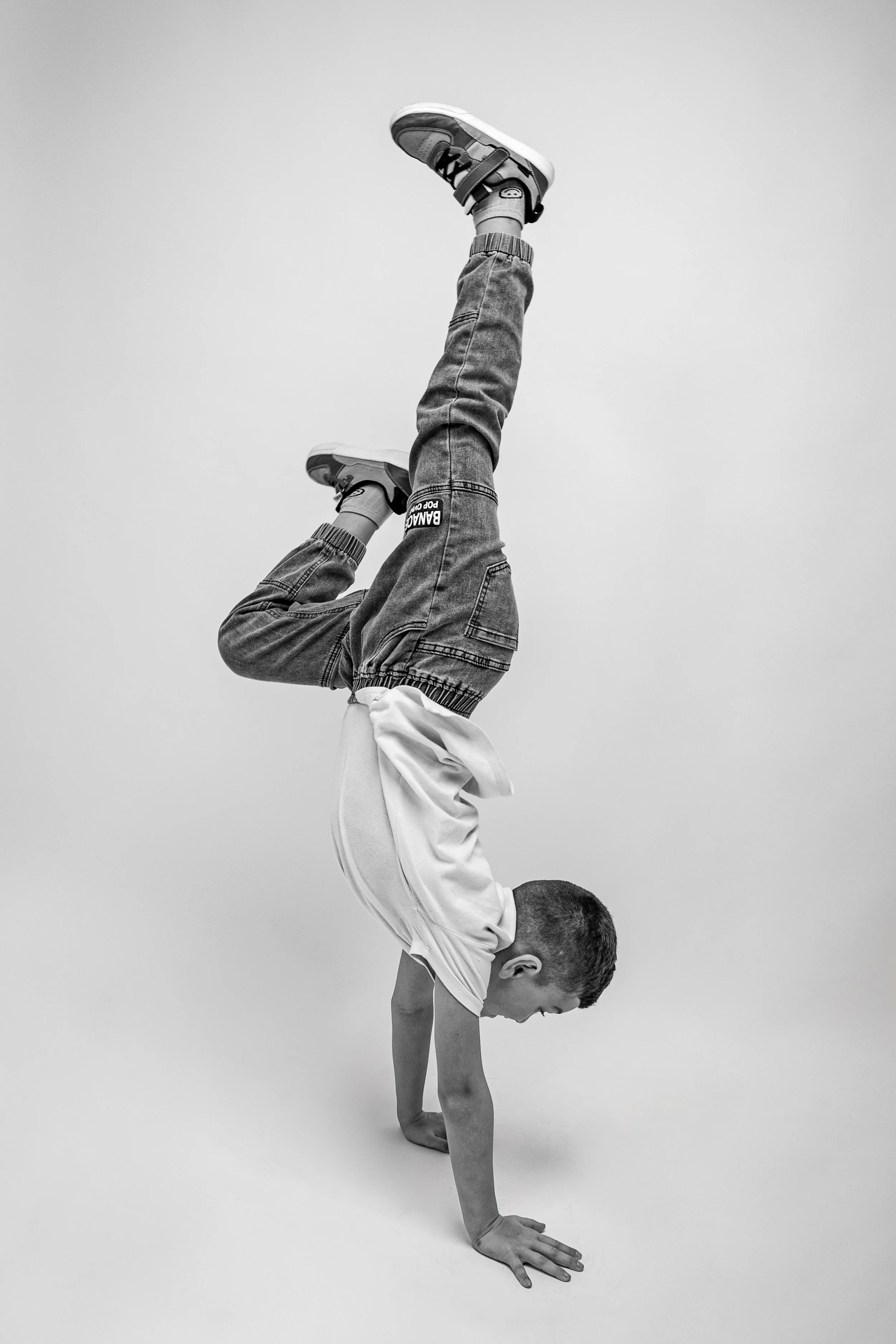 A person performing a handstand, with their legs bent backwards and feet pointed towards the sky, dressed in denim pants, sneakers, and a white shirt, against a plain background.