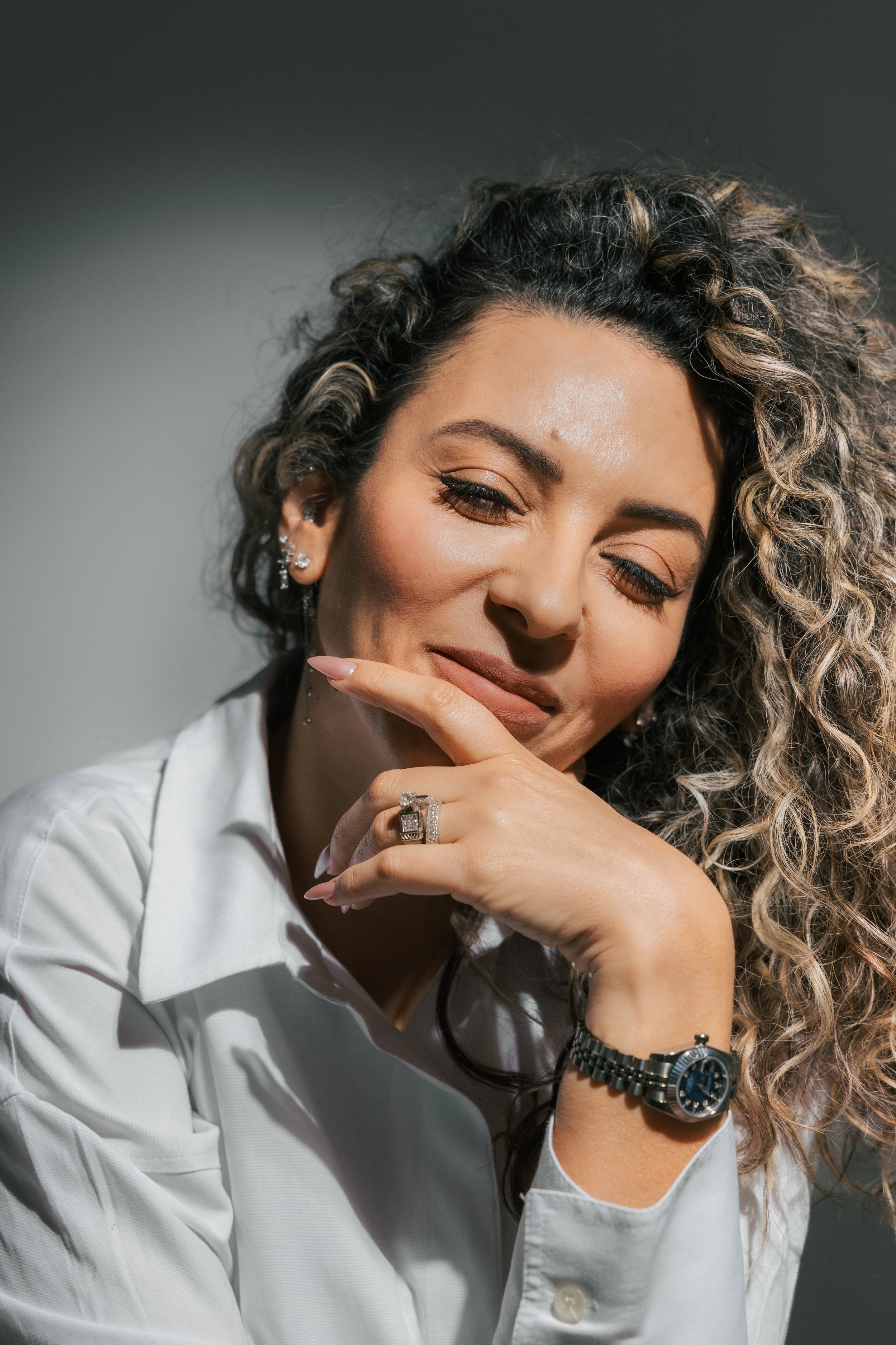 A woman with curly hair, wearing earrings and a watch, smiling and resting her chin on her hand in a white shirt.