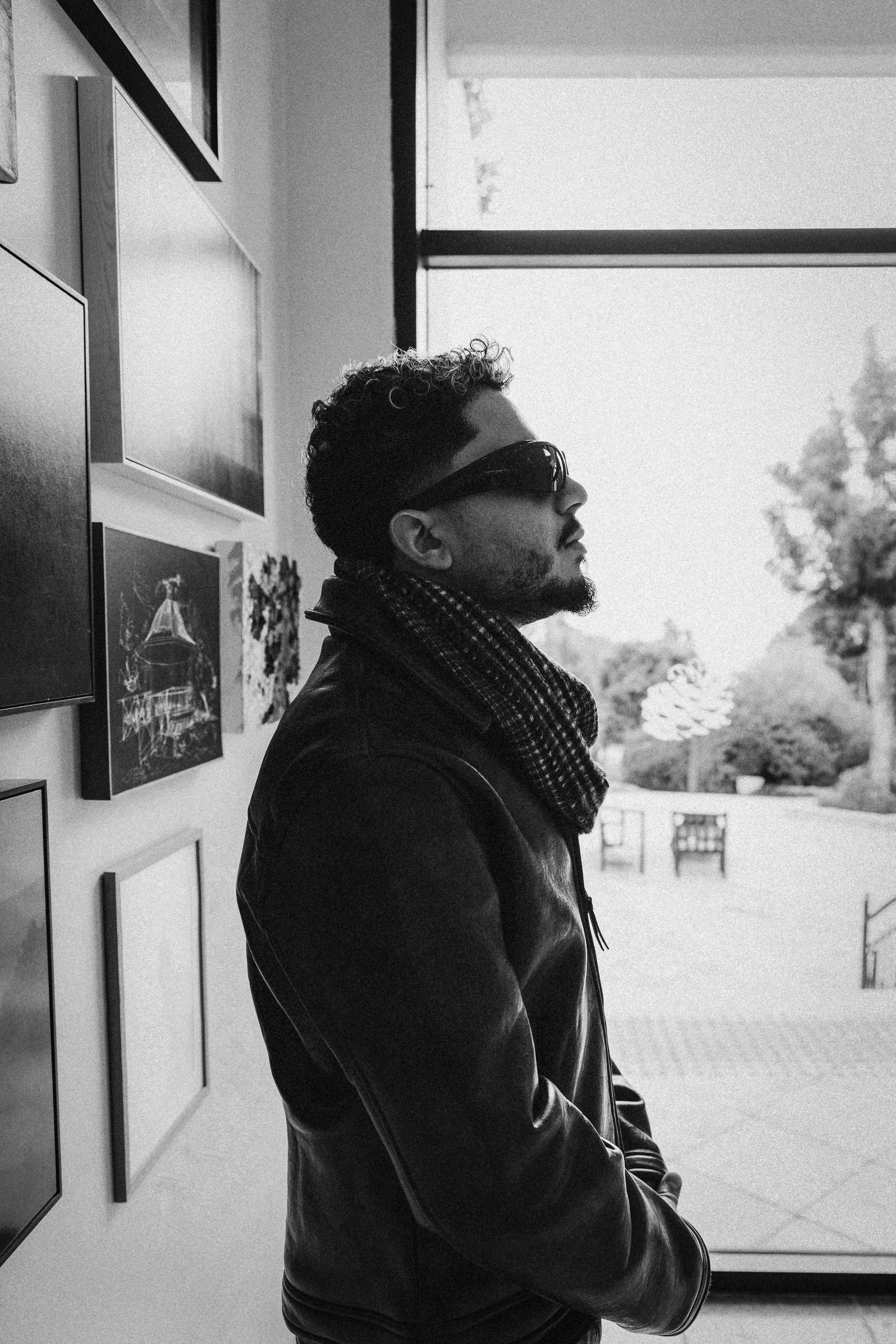 A man with curly hair and a beard wearing sunglasses and a leather jacket stands in profile, looking out a large window. The room has artwork on the walls, and outside trees and a bright sky are visible.