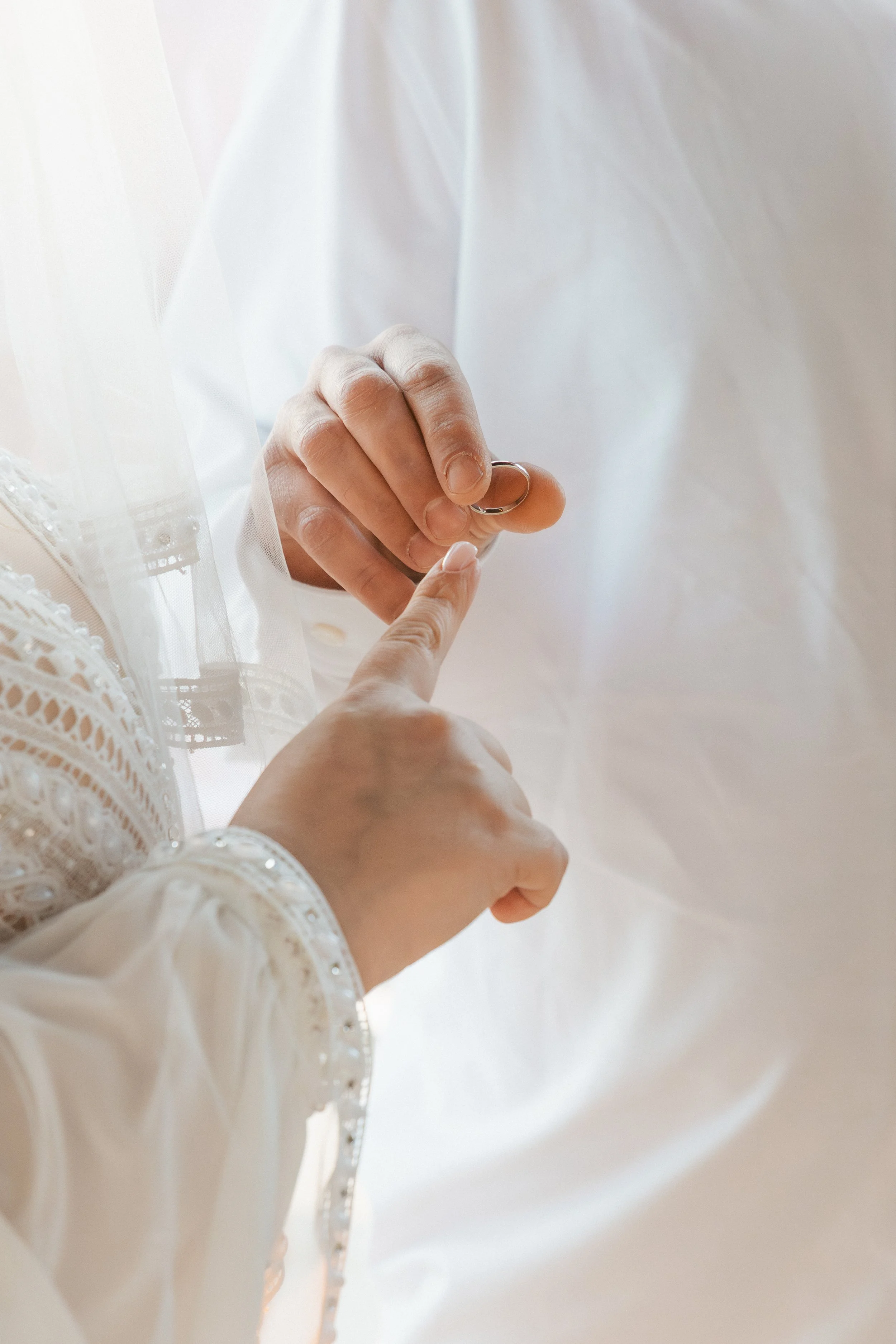 Close-up of a person holding a wedding ring, dressed in a white lace wedding gown, with light shining and creating a soft glow in the background.