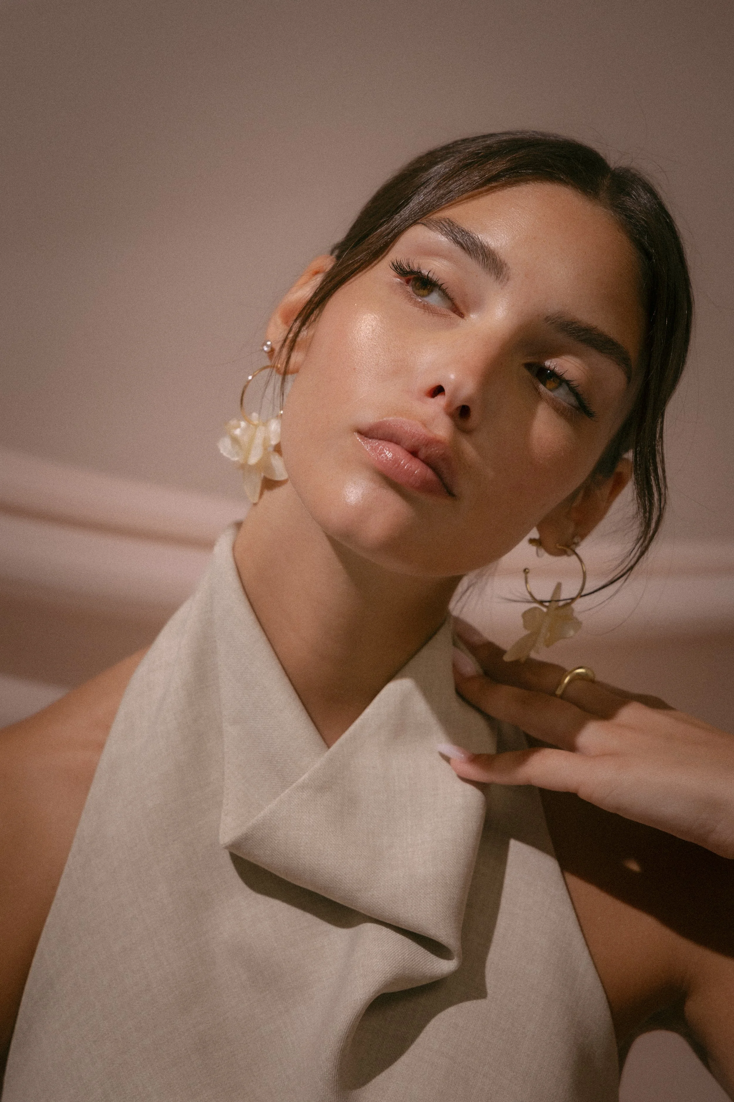 A close-up portrait of a young woman with dark brown hair styled in a sleek manner, wearing large floral earrings, and a beige sleeveless top. She has natural makeup with glossy lips and is gazing to her right with a calm expression against a neutral