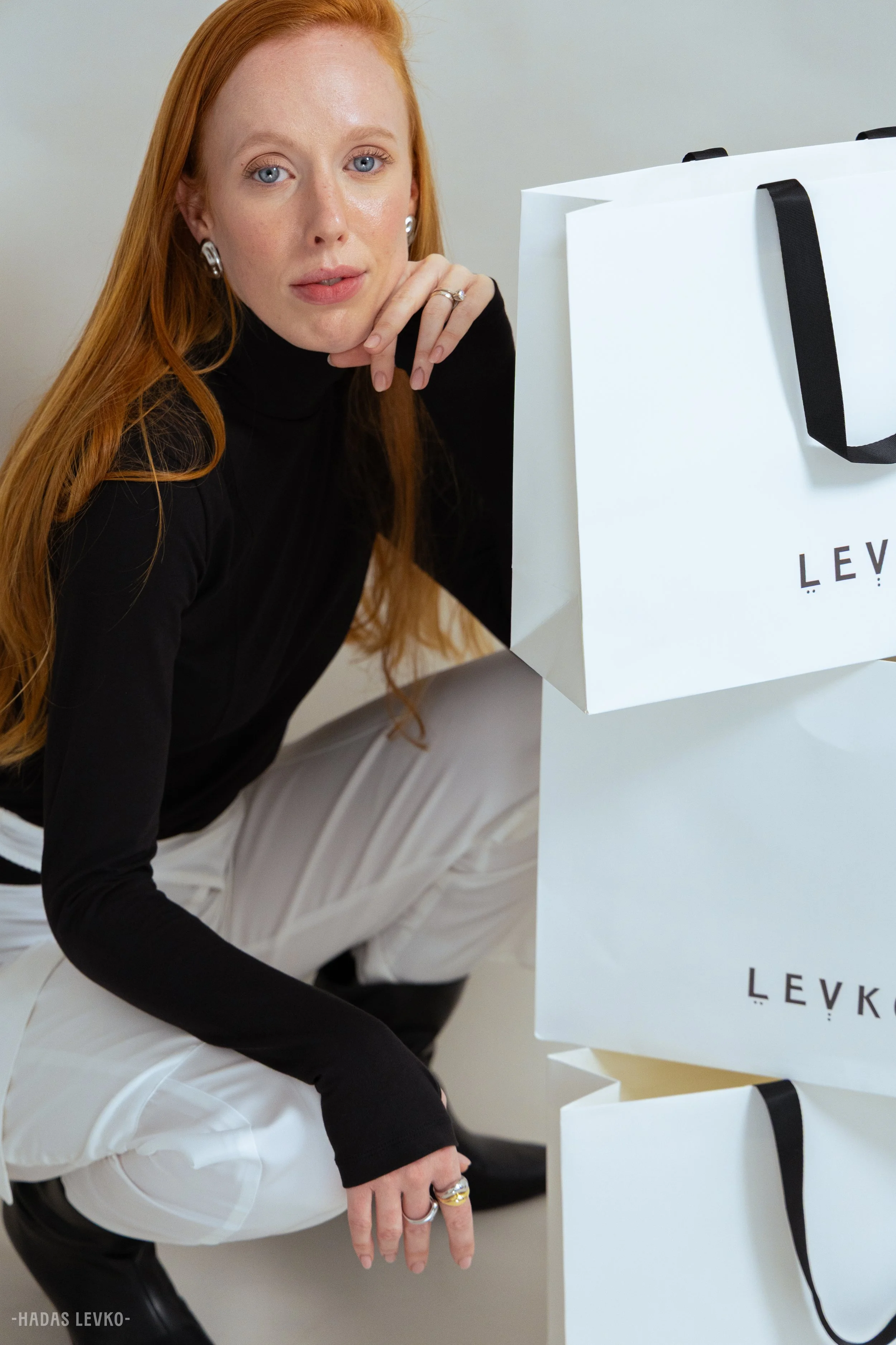 A woman with red hair and blue eyes, wearing a black turtleneck and white pants, sits near white shopping bags with black handles, with her hand resting on her face and her legs crossed.