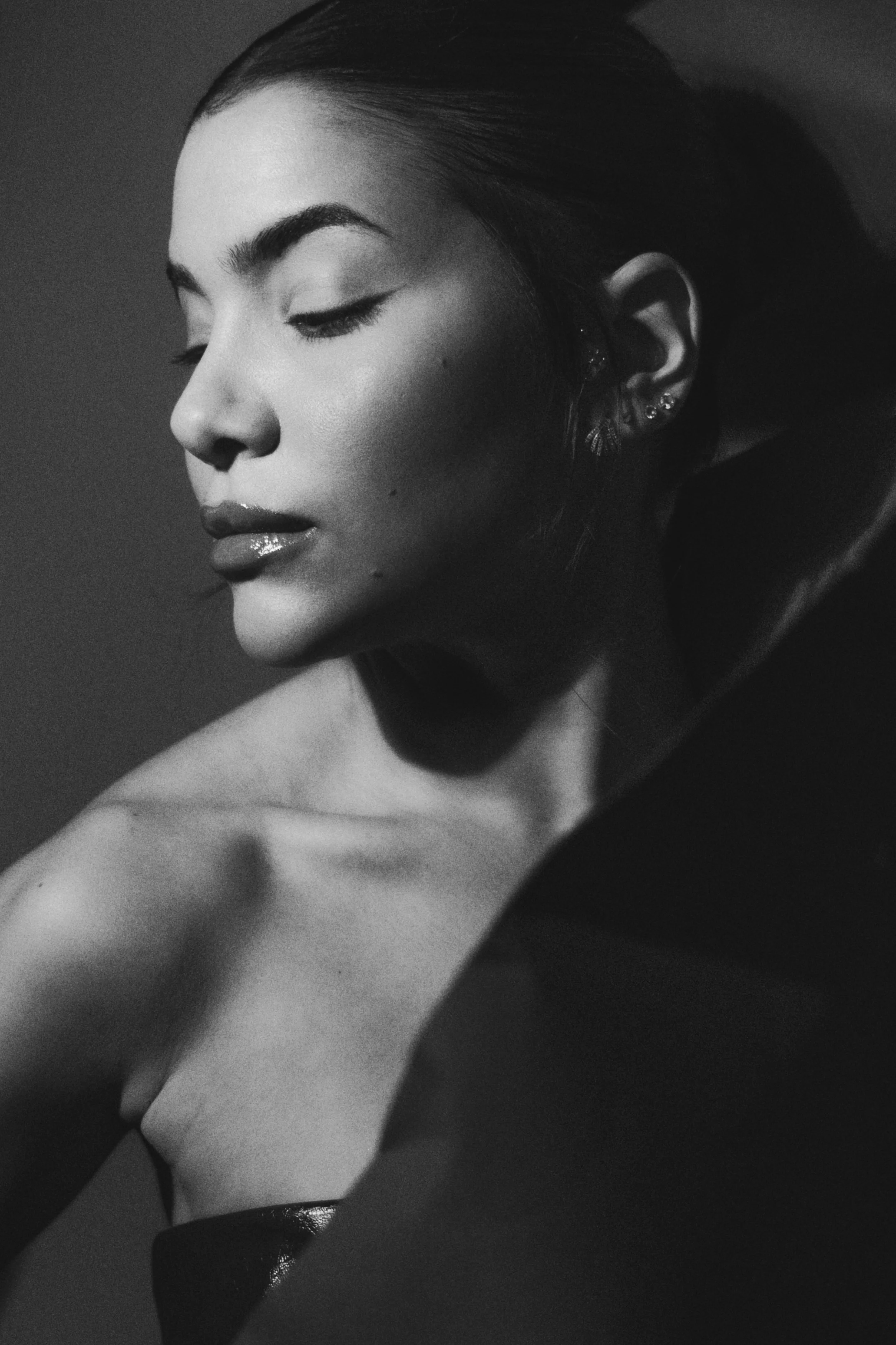 A black and white portrait of a woman with her eyes closed, showing her face from the left side and her shoulder, with multiple earrings in her ear and smooth, styled hair.