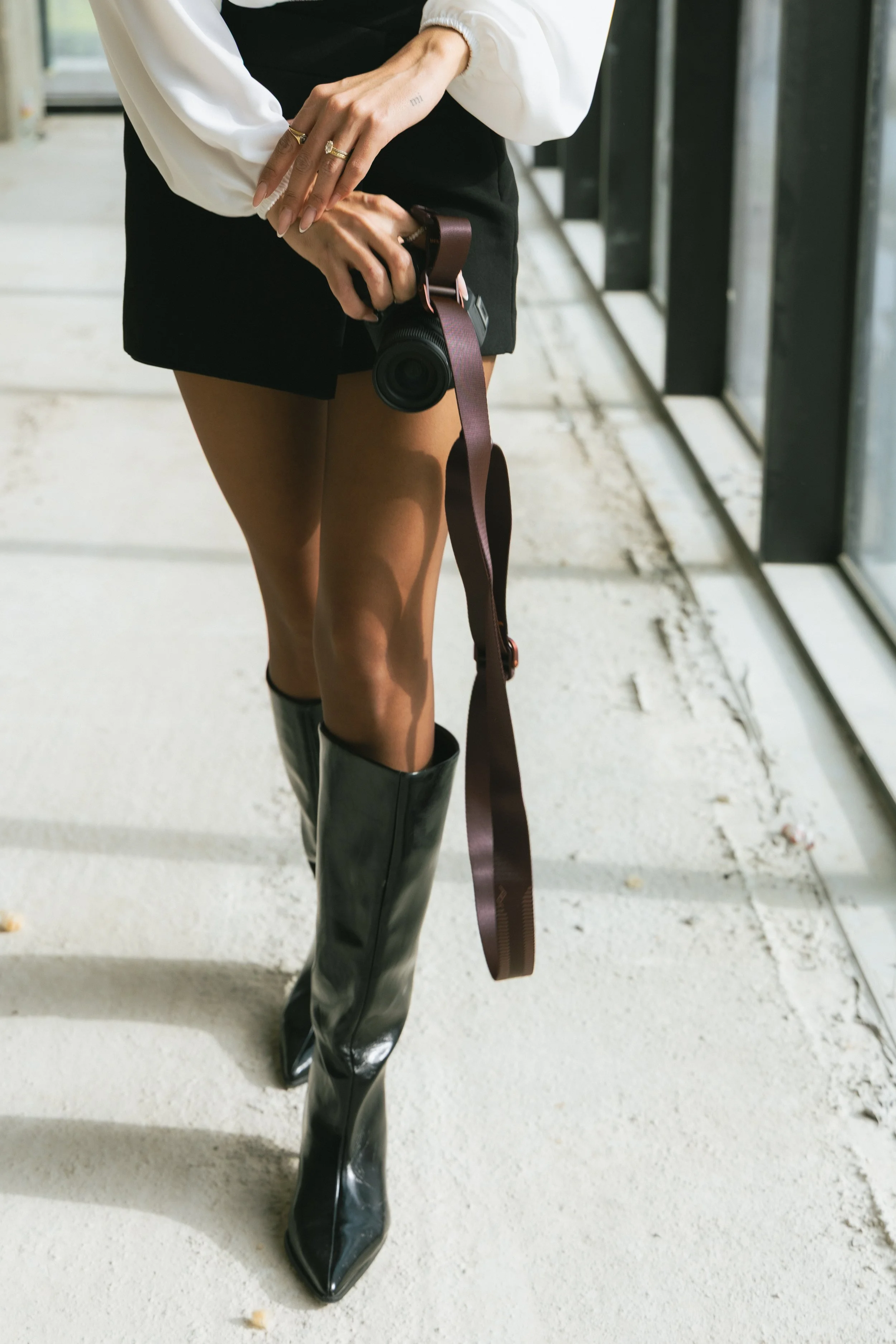 Person wearing black shiny thigh-high boots, black shorts, a white blouse, and holding a camera with a brown strap, standing near large windows with a concrete floor.