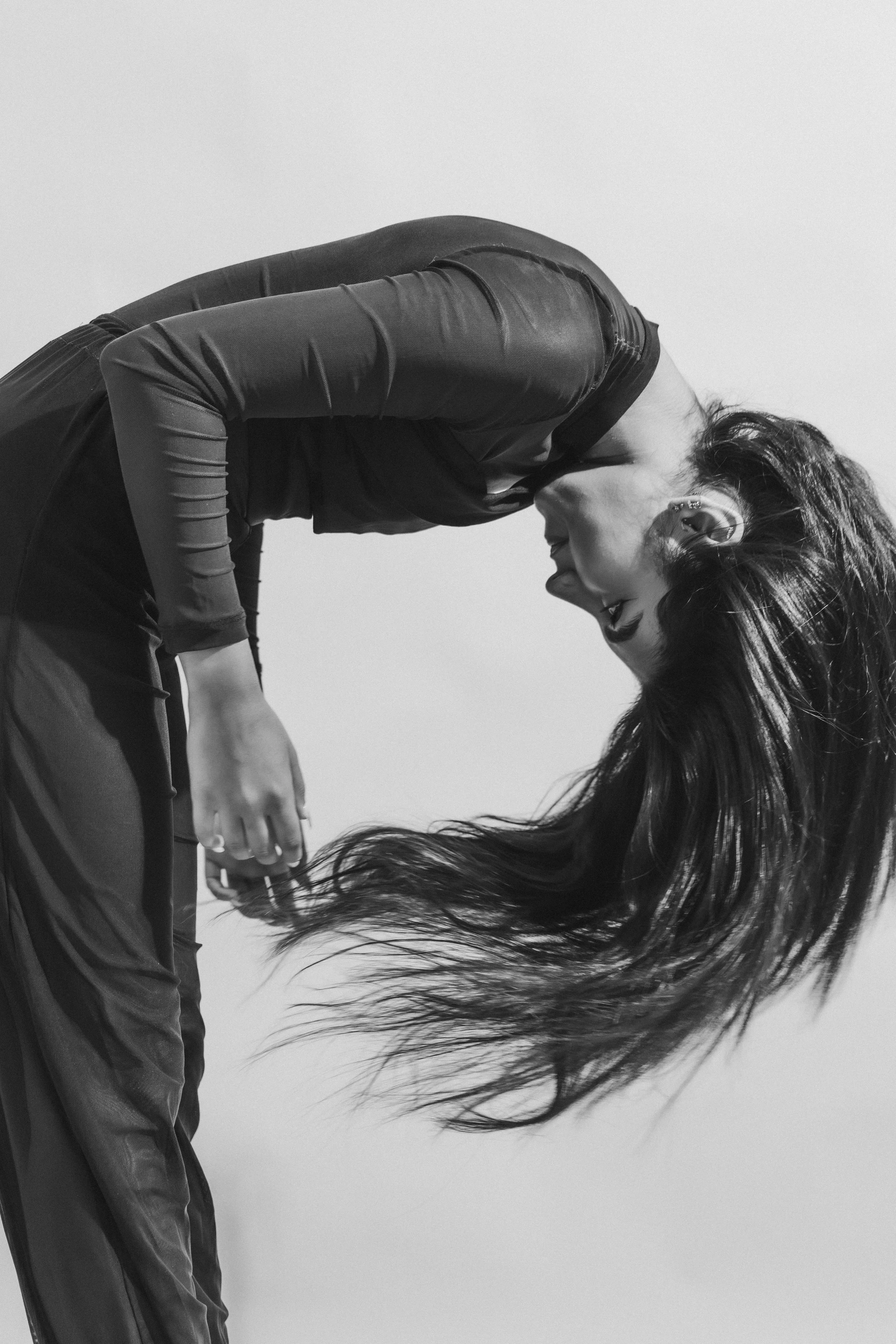 A woman with long, flowing hair leaning forward with her head tilted down. She is dressed in dark athletic clothing, and the background is plain and light-colored.