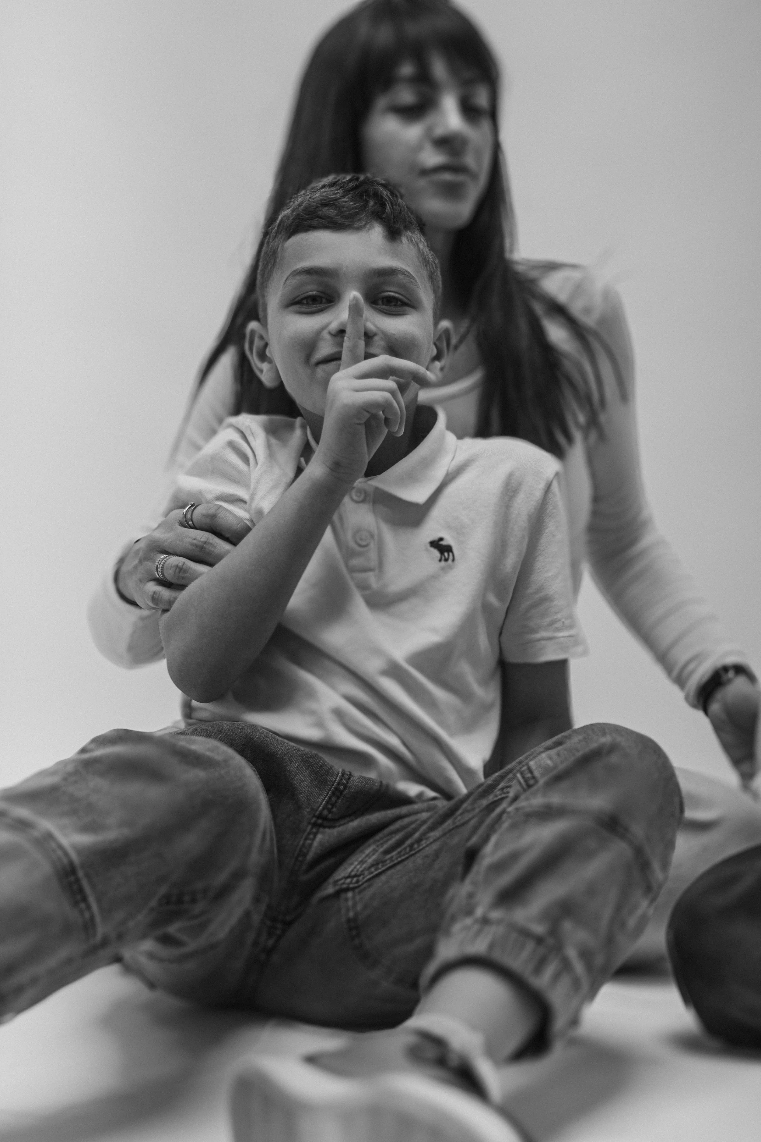 Black and white photo of a young boy sitting on the floor with a woman behind him. The boy is smiling, holding a finger to his lips in a shushing gesture. He is wearing a polo shirt and jeans. The woman, possibly his mother, has long dark hair and is